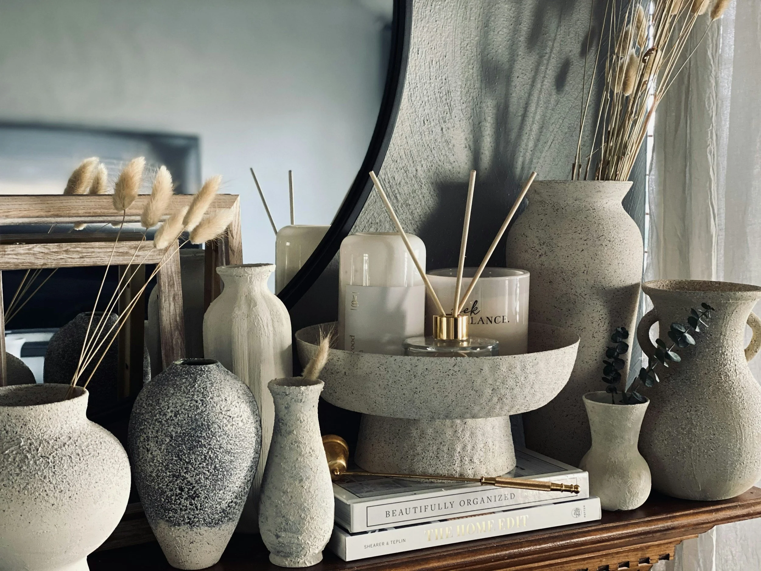 Various vases and jars with dried plants and eucalyptus, set on a wooden surface next to a mirror and a window with curtains.