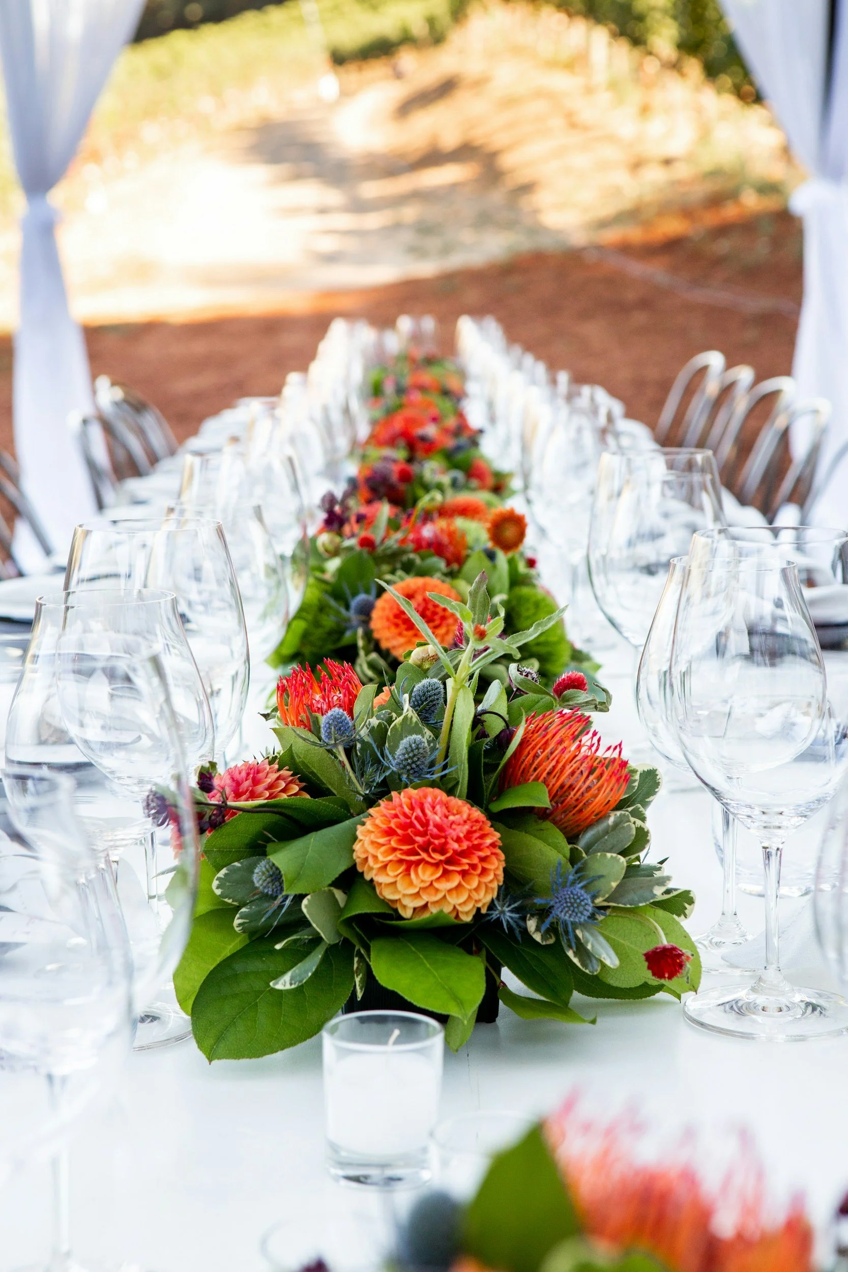 A long outdoor dining table decorated with a vibrant floral centerpiece, surrounded by wine glasses and chairs, set in a natural setting with trees and dirt path in the background.