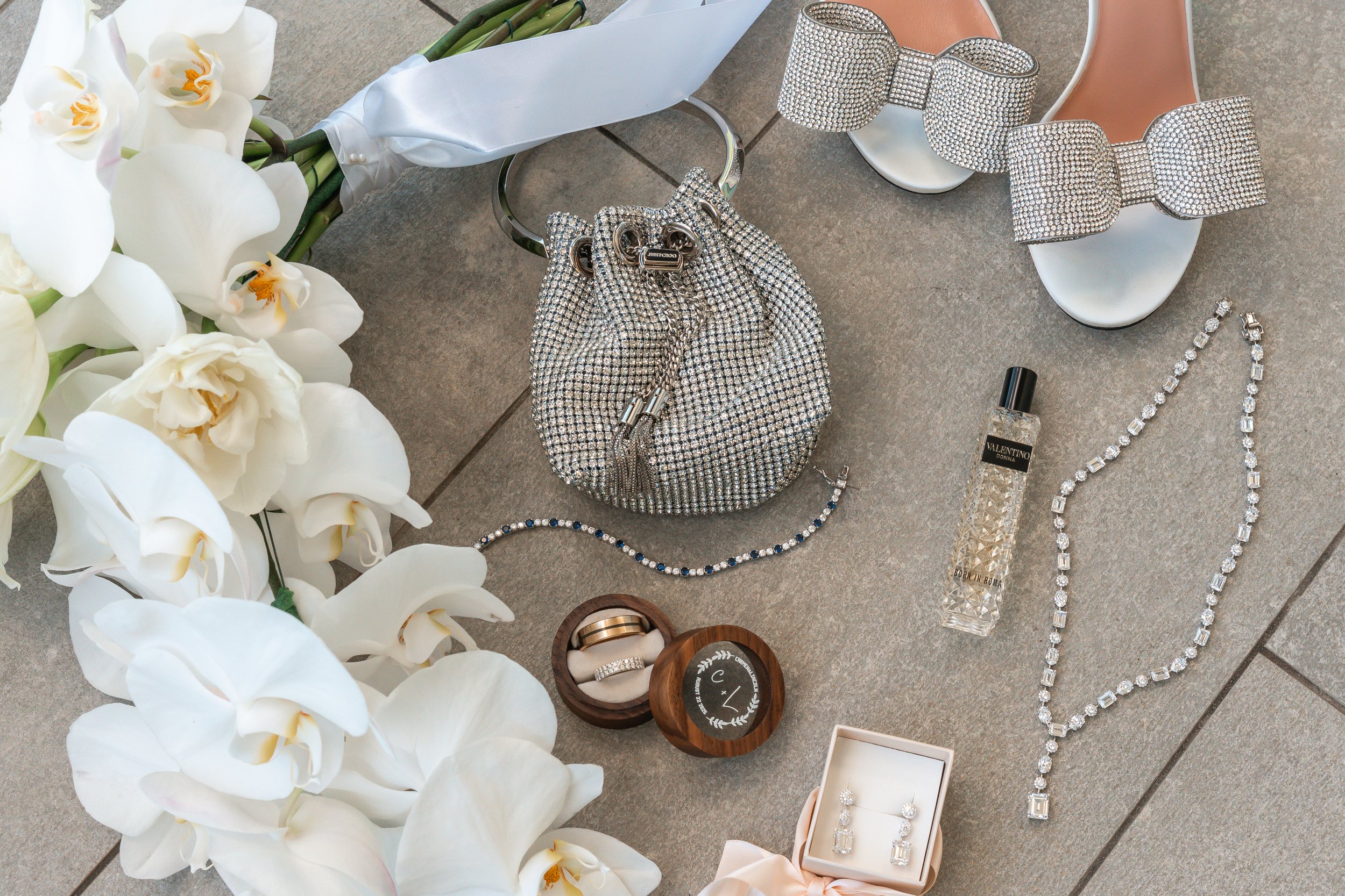 Arranged wedding accessories and jewelry on a tiled floor, including white orchids, diamond bracelets, earrings, necklaces, rings, a small silver purse, and a bottle of perfume.