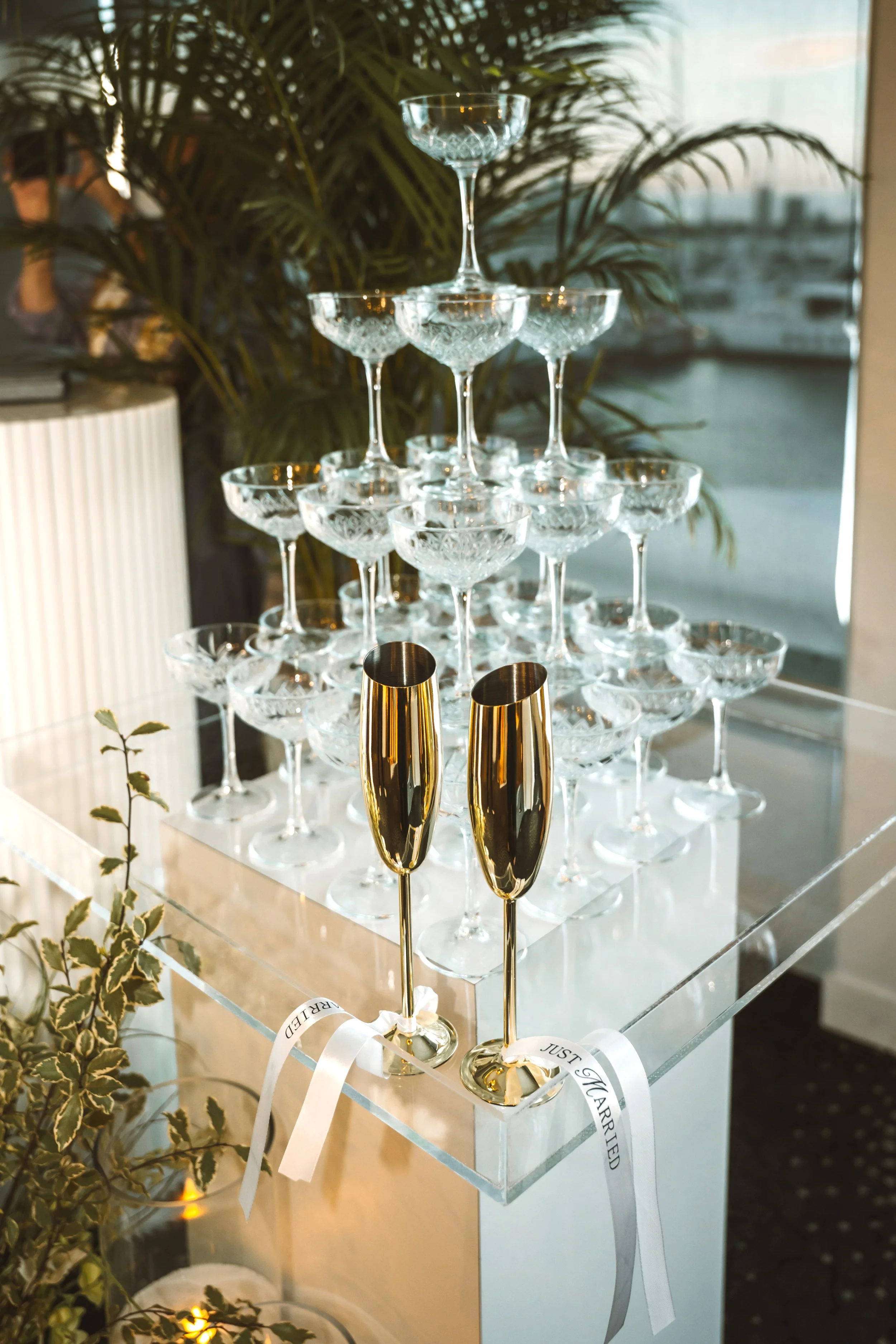 A pyramid of vintage cut glass champagne coupes and two gold champagne flutes on a clear acrylic table, with decorative greenery and a harbor view in the background.