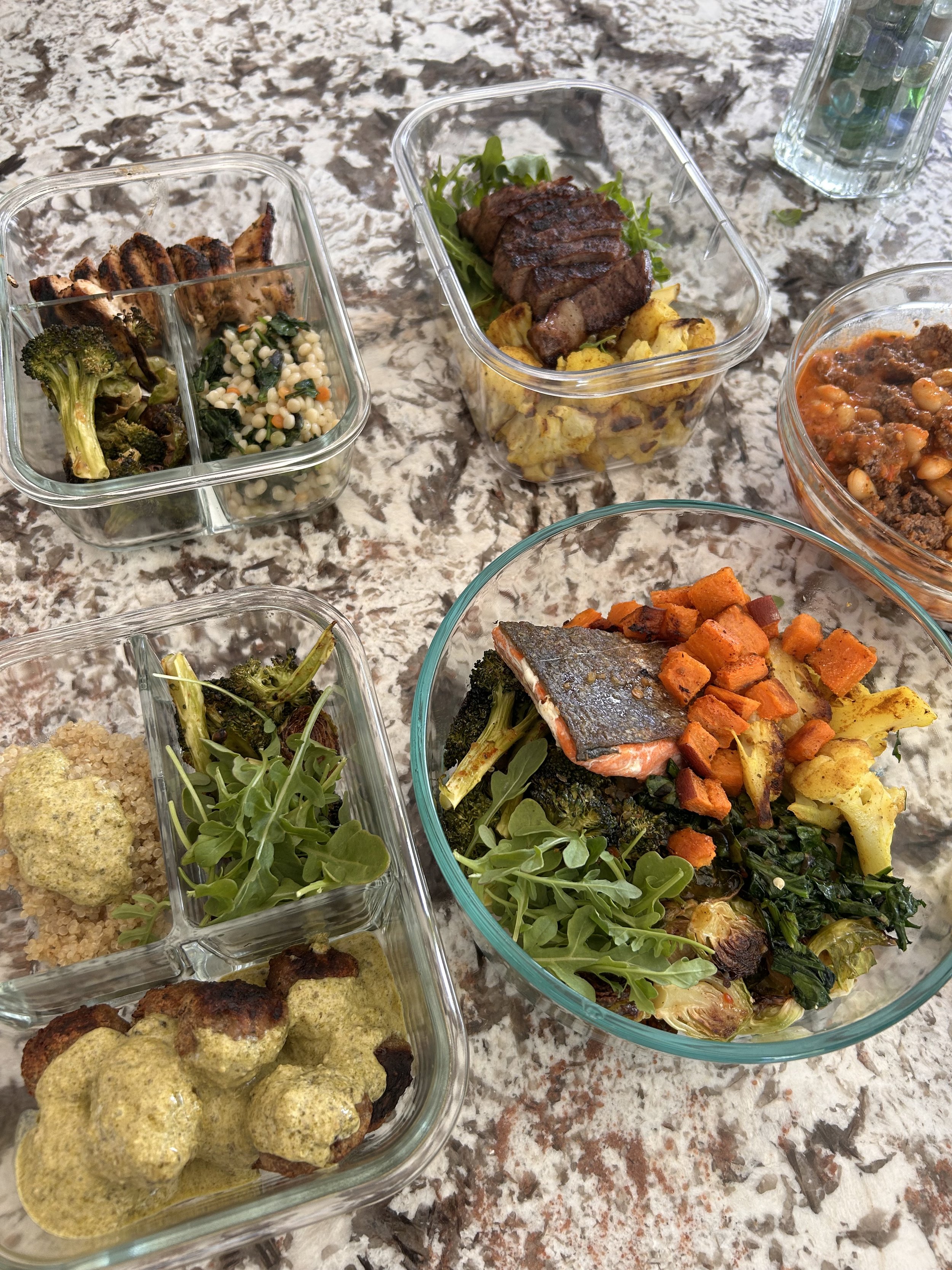 Assorted dishes of grilled meats, roasted vegetables, salad, and baked salmon on a marble countertop.
