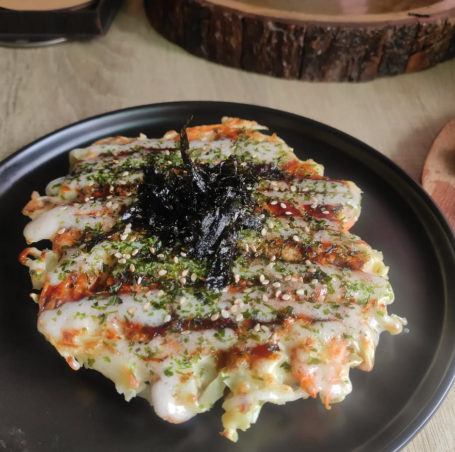 A plate of savory Korean pancake topped with seaweed strips, sesame seeds, and drizzled with sauce.