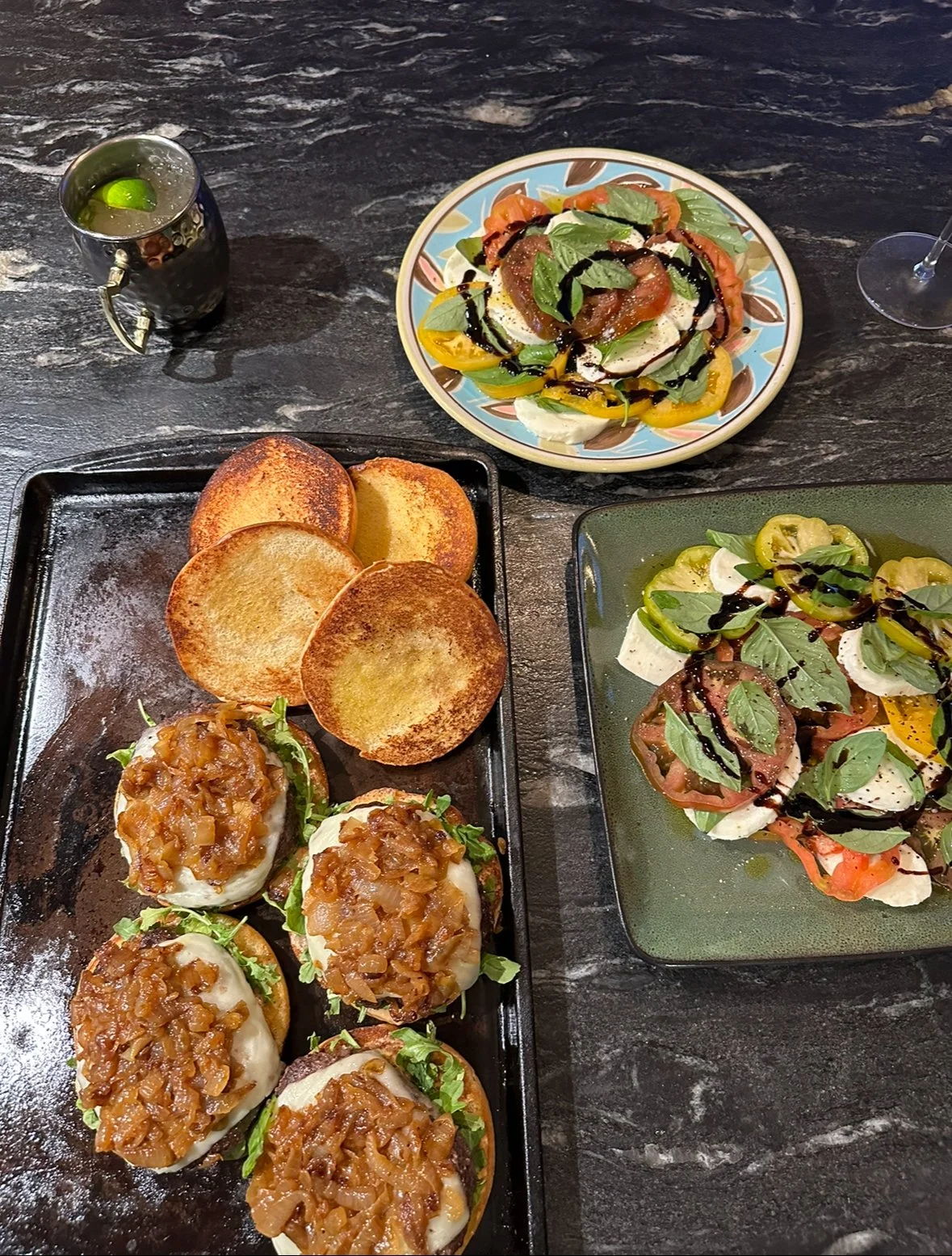 A table with toasted bread, eggs topped with onion relish, and plates of Caprese salad with tomatoes, mozzarella, basil, balsamic glaze, and olive oil, along with a beverage with a lime wedge.