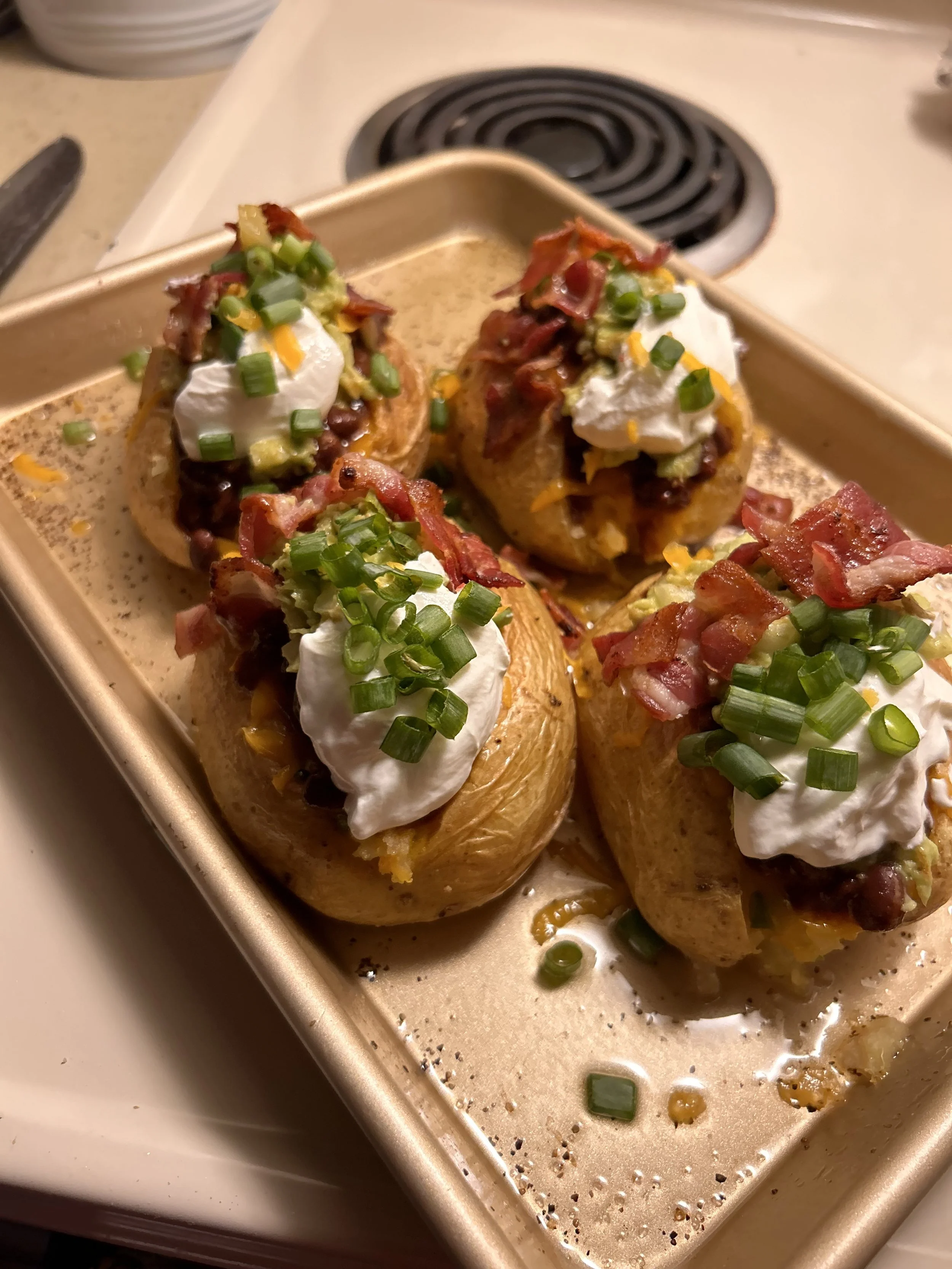 Baked potatoes topped with sour cream, bacon bits, chopped green onions, and shredded cheddar cheese on a baking tray.