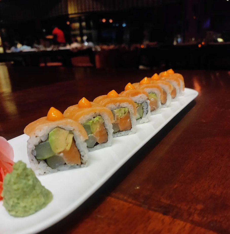 A plate of sushi rolls with avocado, salmon, and cream cheese, topped with orange sauce dots on a white rectangular dish, with wasabi and ginger on the side, in a dimly lit restaurant setting.