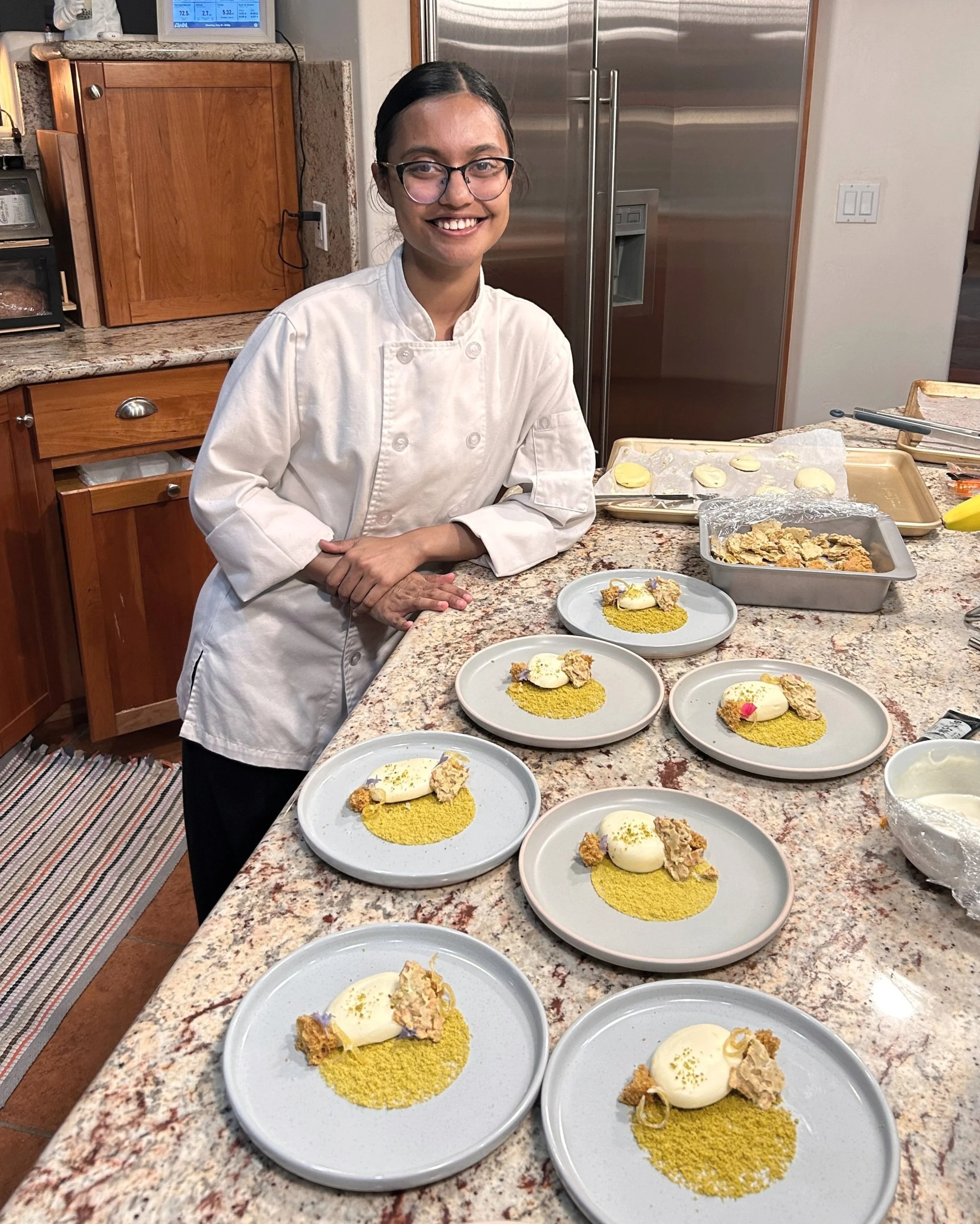 A woman in a chef's coat stands in a kitchen with a wide smile, surrounded by plates of food on a granite countertop. The plates feature a yellow powder, a white scoop of ice cream or similar, and crumbled toppings, with some garnished with small colorful decorations. There are baked goods and cooking utensils in the background.