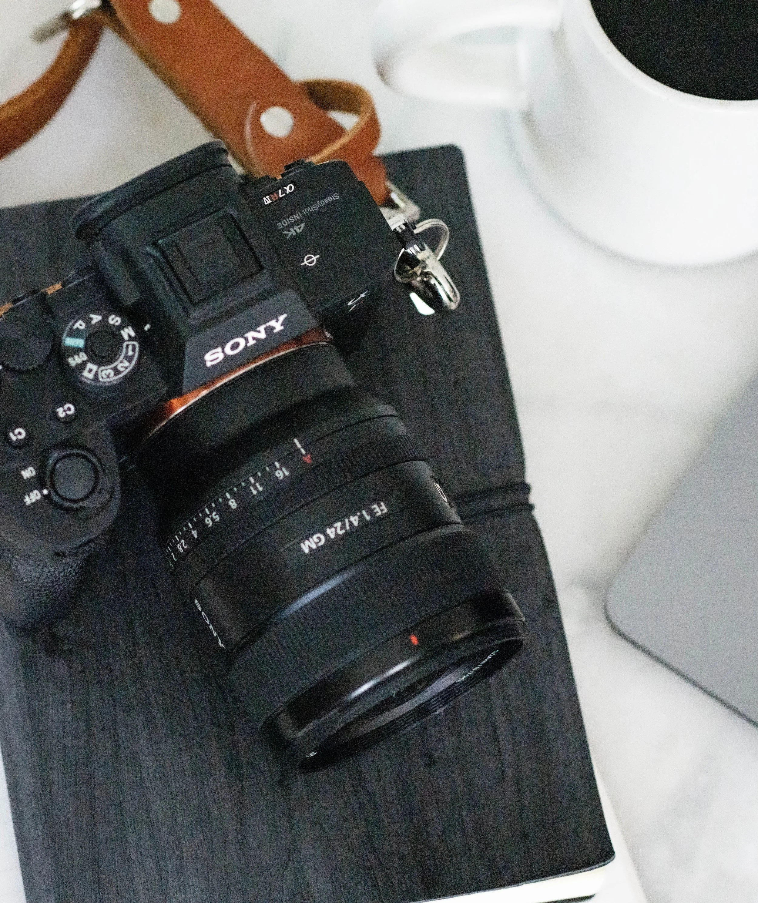 Close-up of a black Sony digital camera with a zoom lens, placed on a flat surface next to a white mug and a brown leather strap.
