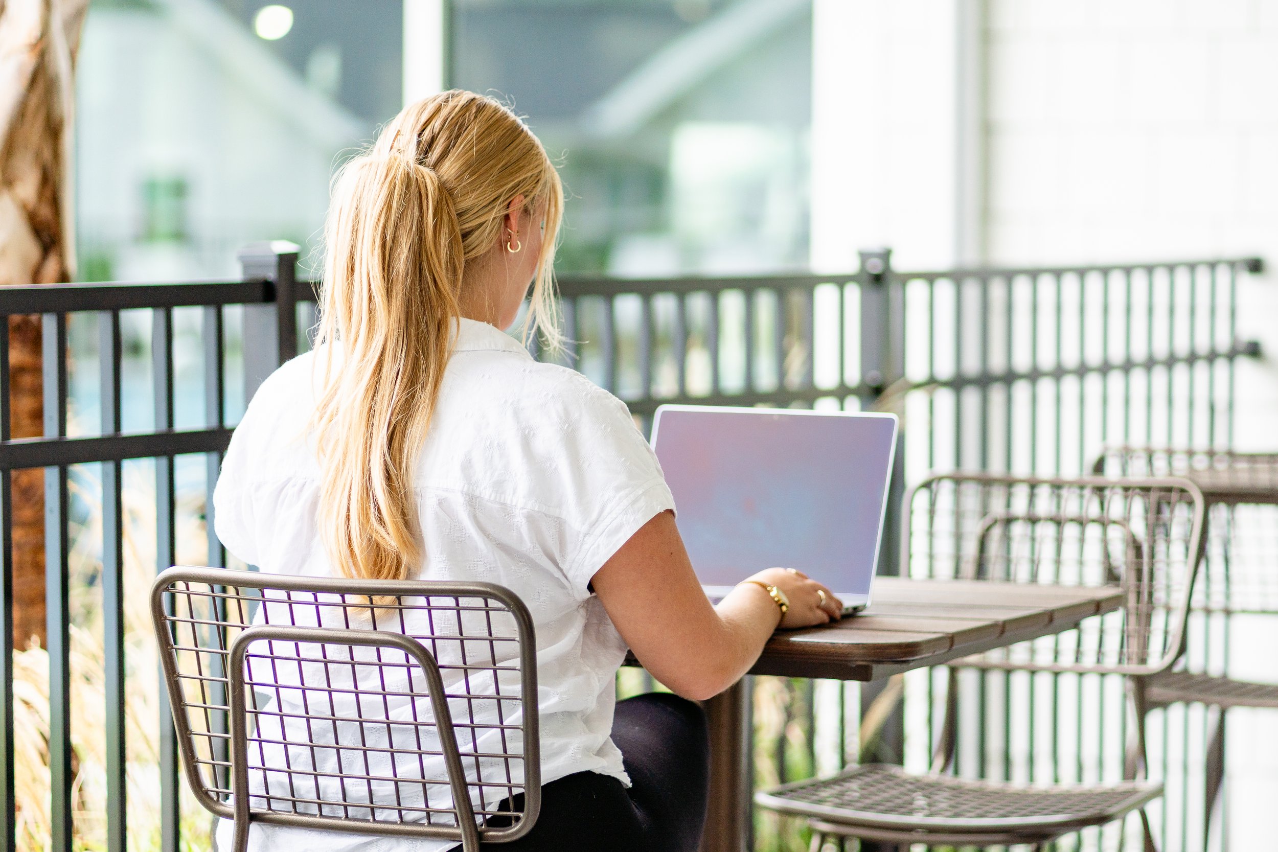 A woman with long blonde hair sitting at an outdoor table with a laptop, wearing a white blouse and gold jewelry, in a bright, modern setting with glass walls.