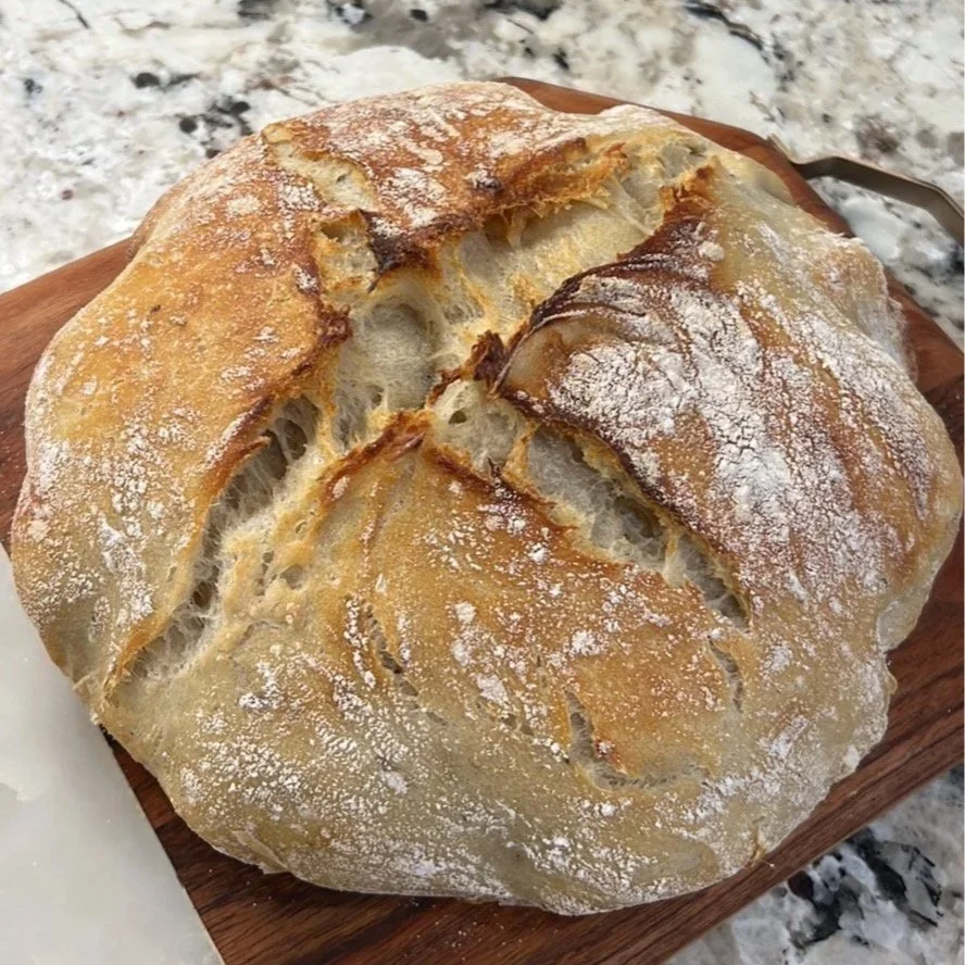 A fresh baked loaf of sourdough bread with a cracked crust, dusted with flour, resting on a wooden cutting board.