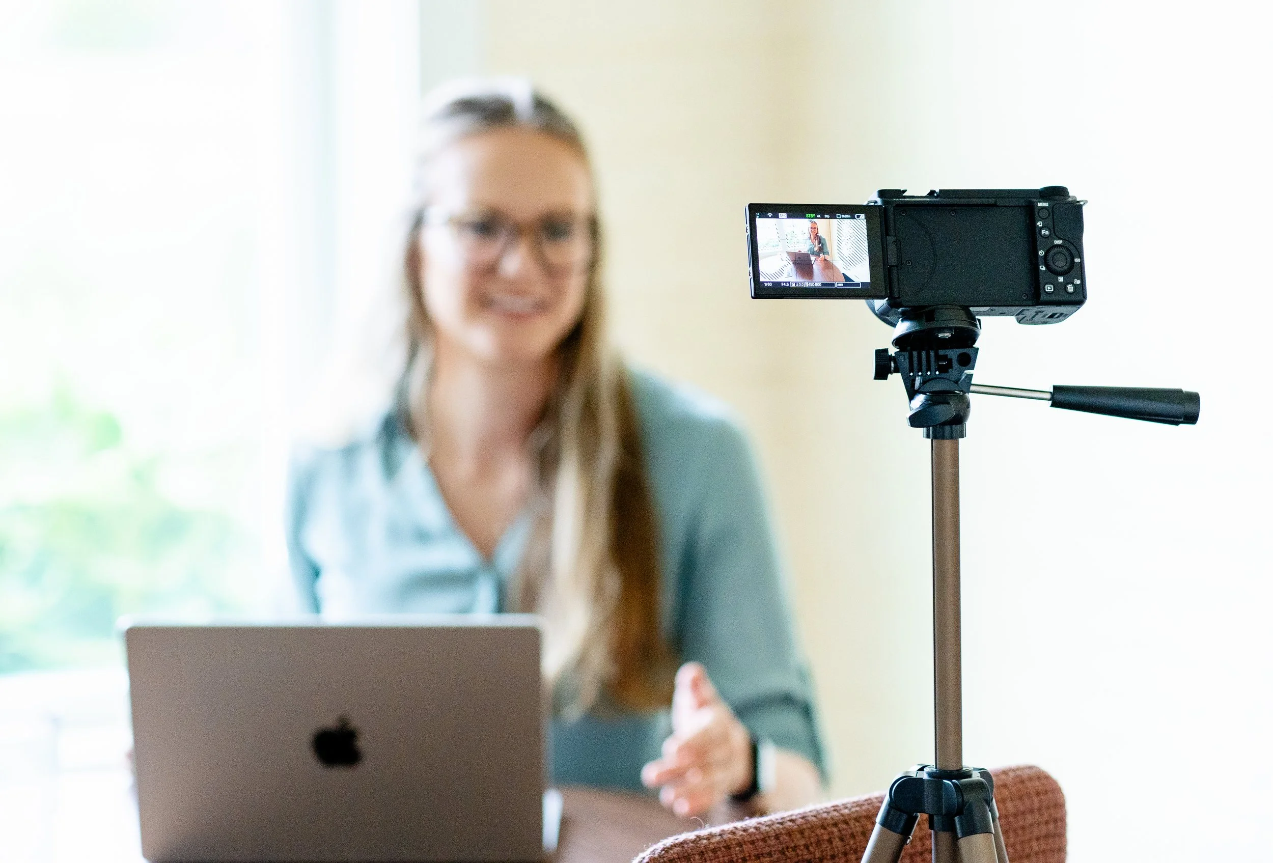 Kelly in glasses working at her laptop, with a camera on a tripod set up to film her