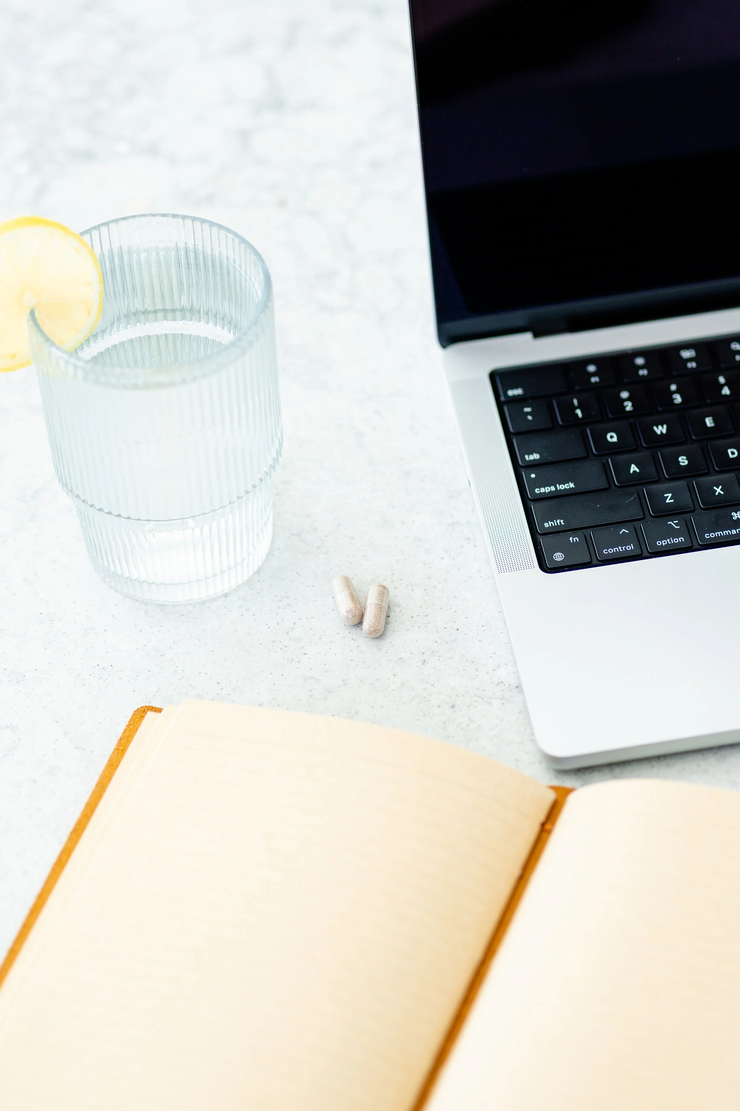 A glass of water with lemon slices, a laptop keyboard, two capsules, an open book, and a closed notebook on a white surface.