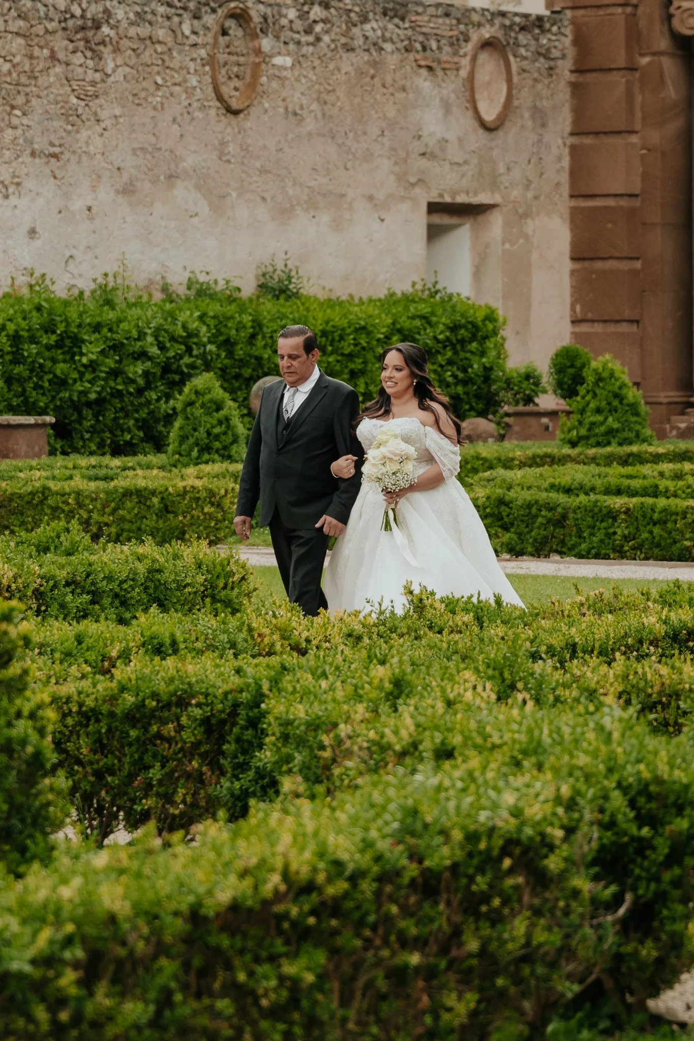 A bride walking outside with a man on her arm, holding a bouquet, in a garden with trimmed bushes and a historic stone building in the background. Rome Italy is the location of the venue.
