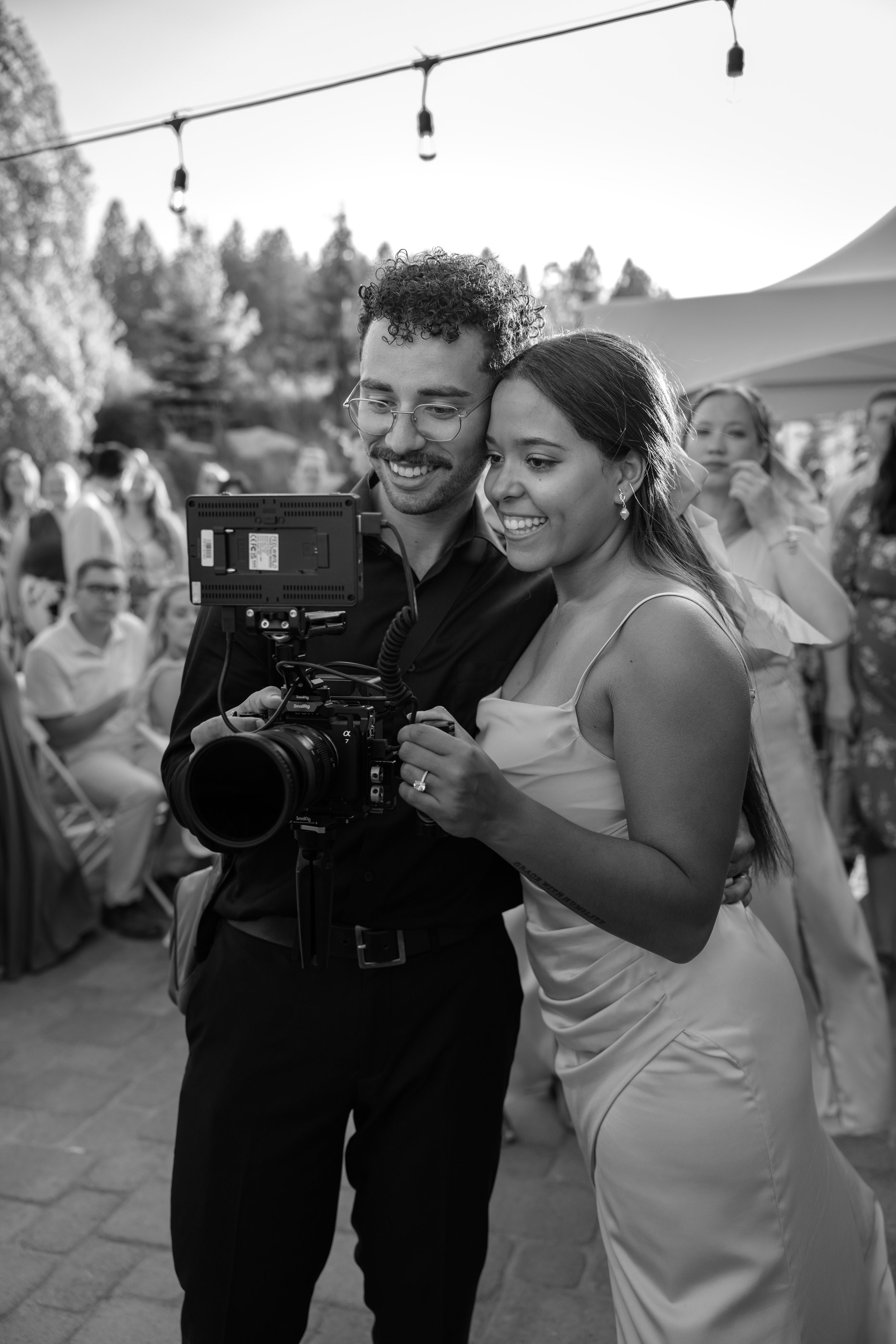 Two people, a man with glasses and a woman in a dress, look at a camera at an outdoor event with people in the background.
