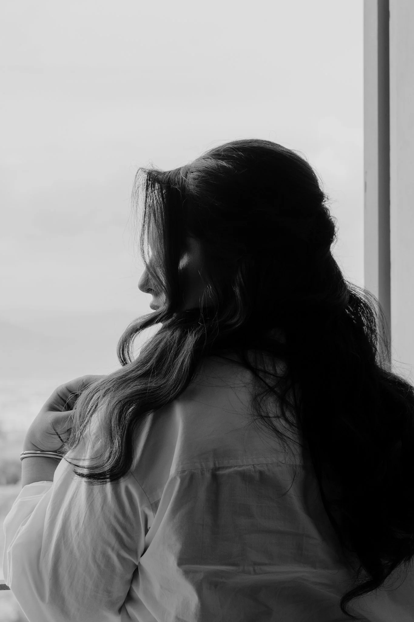 Black and white photo of a woman with long wavy hair, looking out a window, with a cityscape or landscape of Rome in the background.