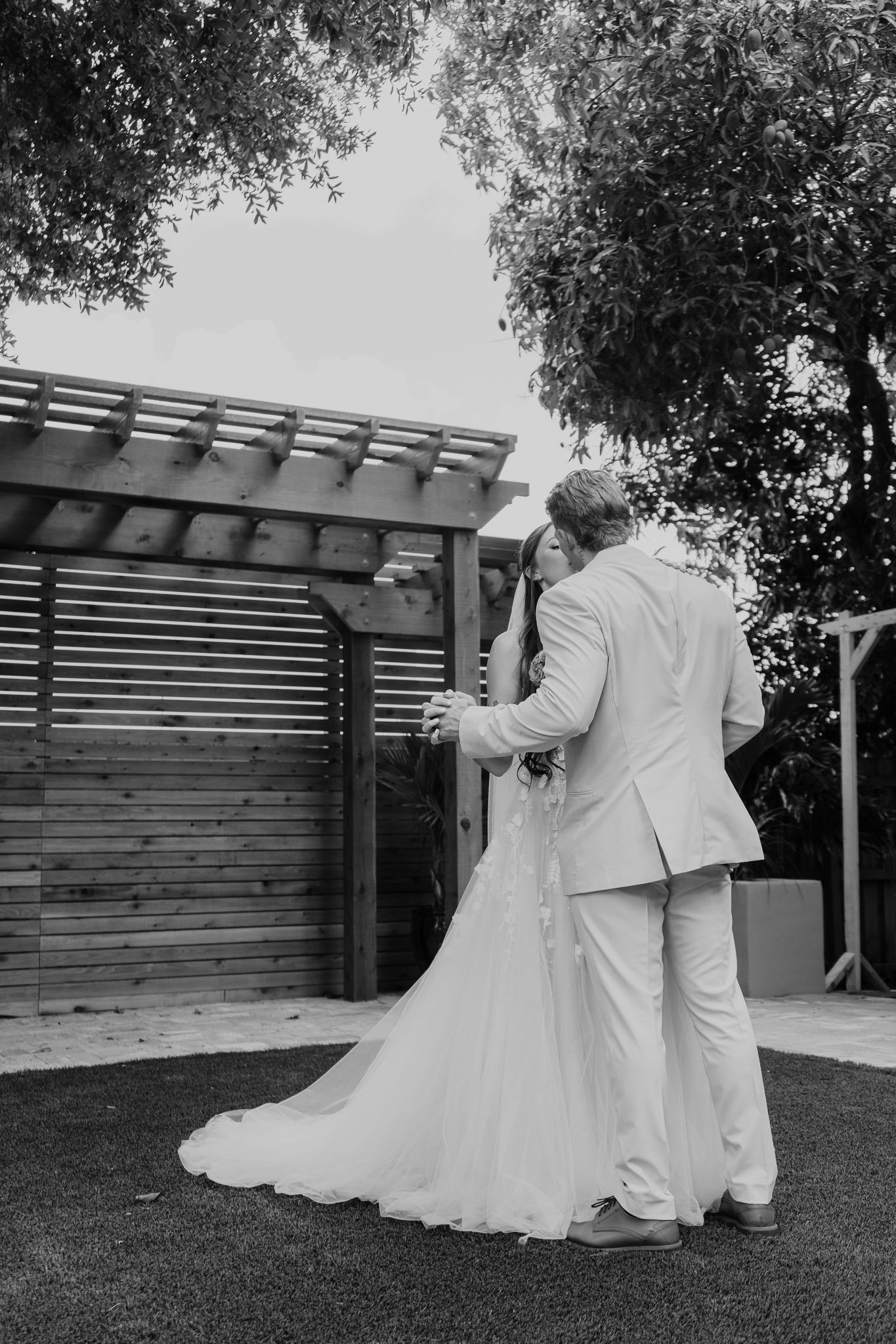 A couple dressed in wedding attire sharing a kiss outdoors, with trees and a wooden structure in the background.