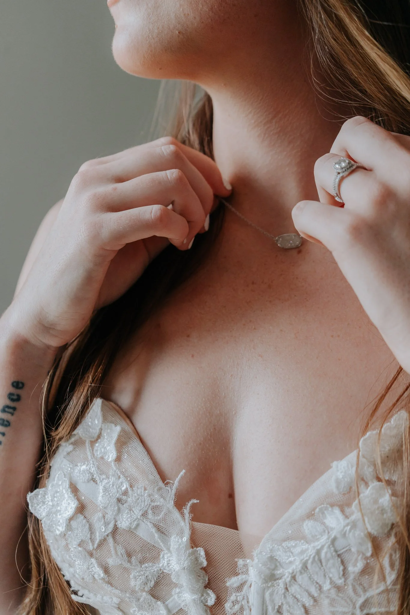 A woman adjusts her necklace, wearing a ring and a lace dress, with a tattoo on her arm. Wedding prep in Florida State.
