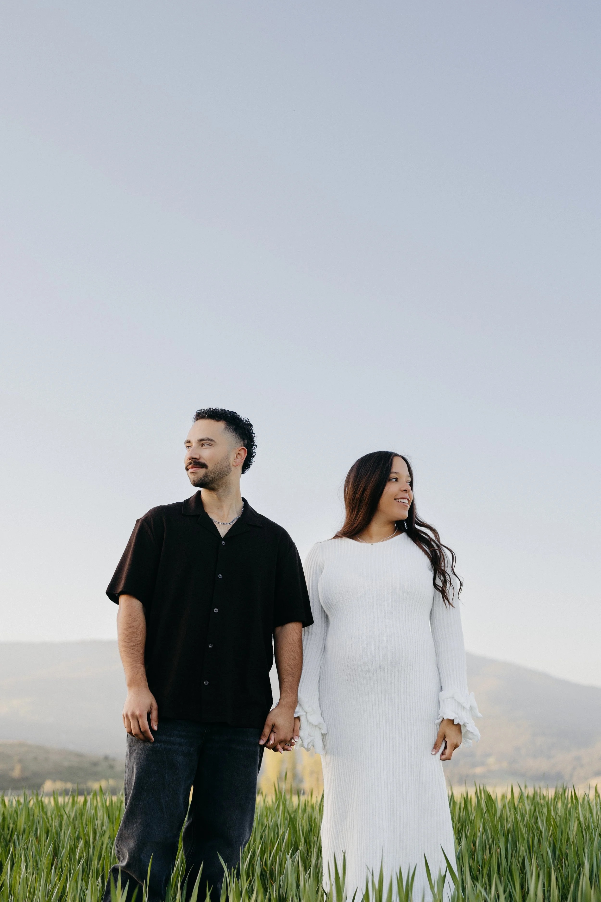 A young man and woman holding hands standing in a grassy field with a cloudy sky in the background.
