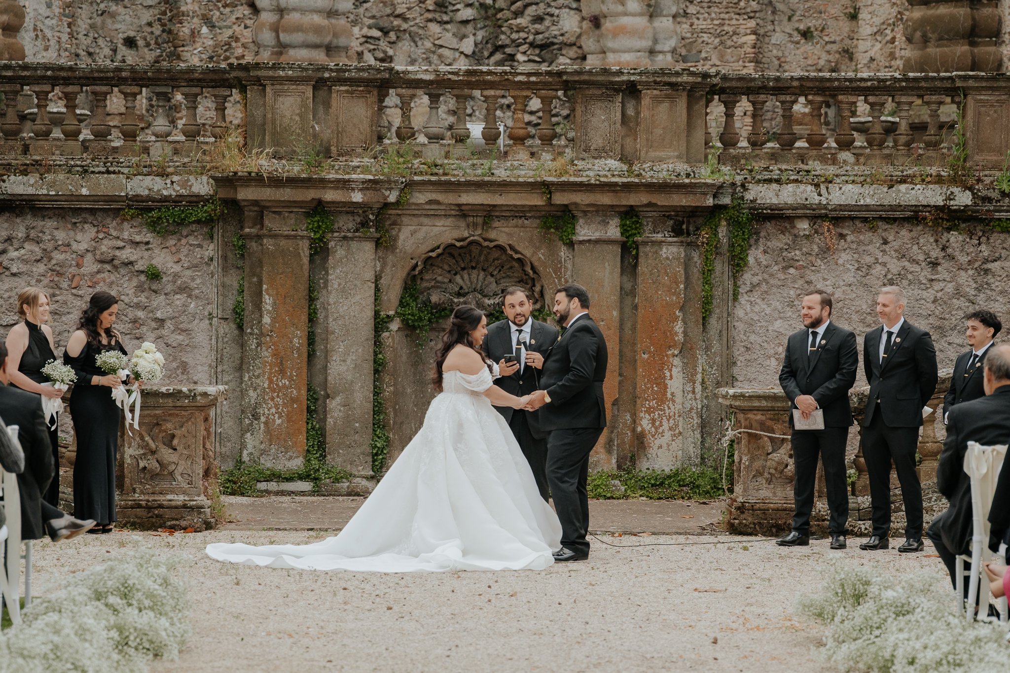 A wedding ceremony taking place outdoors with the bride and groom holding hands, facing each other, in front of a stone backdrop. The bride is wearing a white wedding gown, and the groom is in a black suit. Bridesmaids and groomsmen are standing nearby, dressed in formal black attire. Guests are seated on either side, witnessing the ceremony.