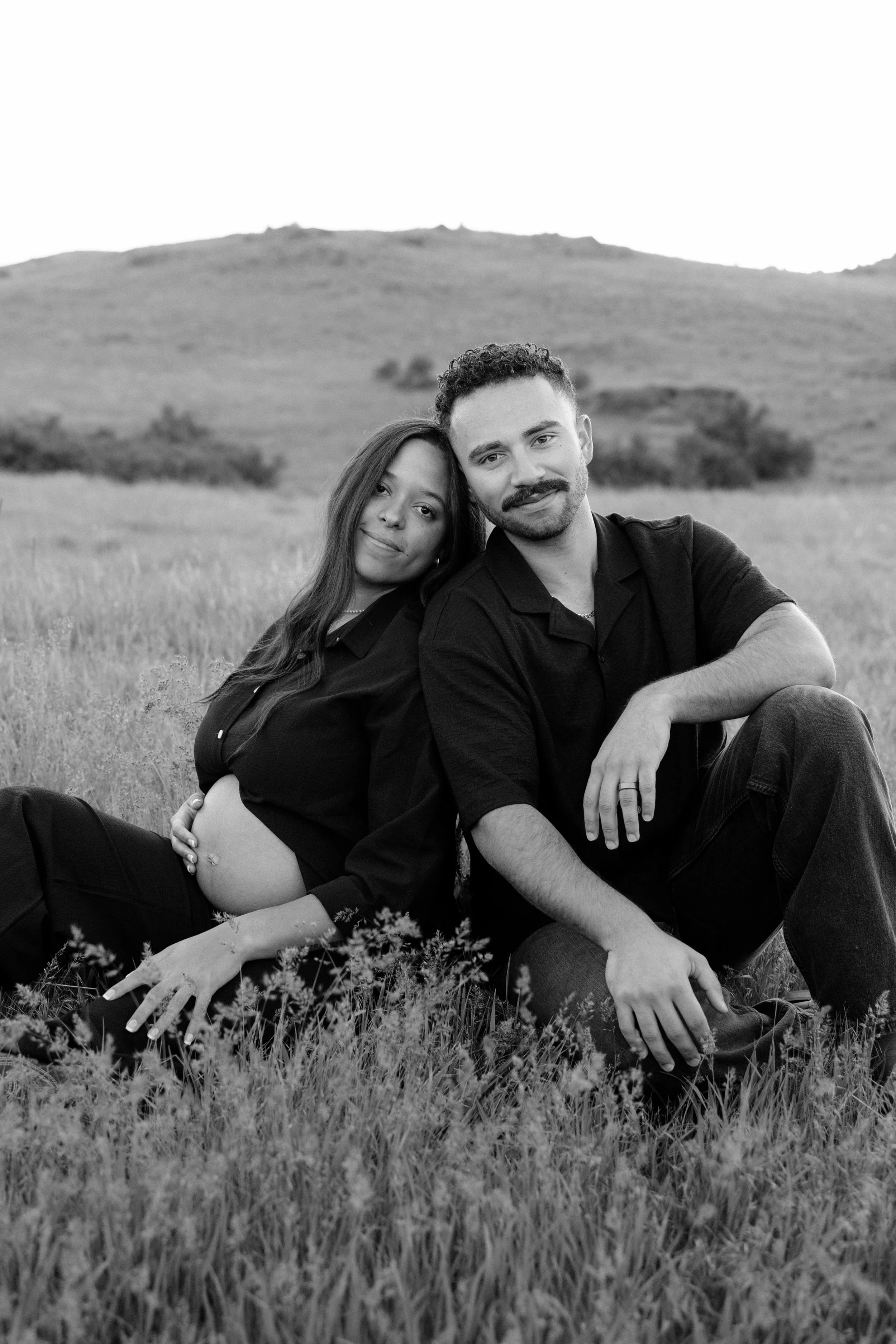 A black and white photo of a couple sitting in a grassy field. The woman has long hair and is leaning her head on the man's shoulder, both smiling. The man has curly hair and a mustache, wearing a dark shirt. Hills are visible in the background.