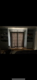 Living room with window covered by curtains, with built-in shelves on either side filled with various items, and a chair in the corner.