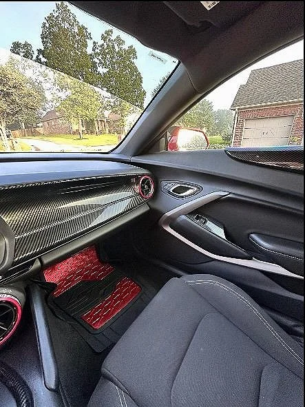 Inside a car, showing a dashboard with a carbon fiber design and red accents, including a red air vent and matching red floor mats. The passenger seat is visible, and outside the window, there is a residential neighborhood with houses and trees.