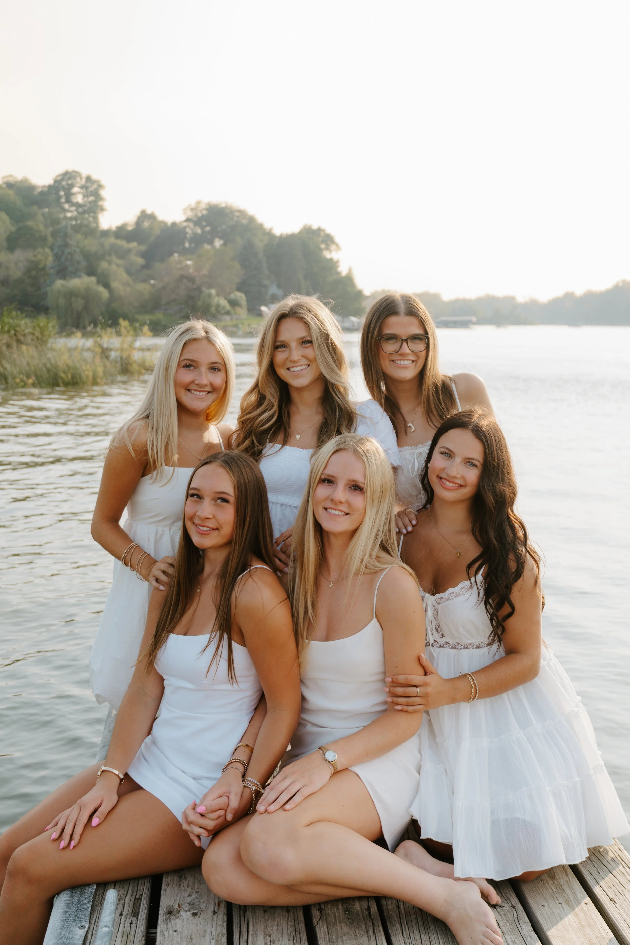 A group of seven young women dressed in white dresses, sitting and standing on a wooden dock by a lake, smiling during sunset with trees and water in the background.