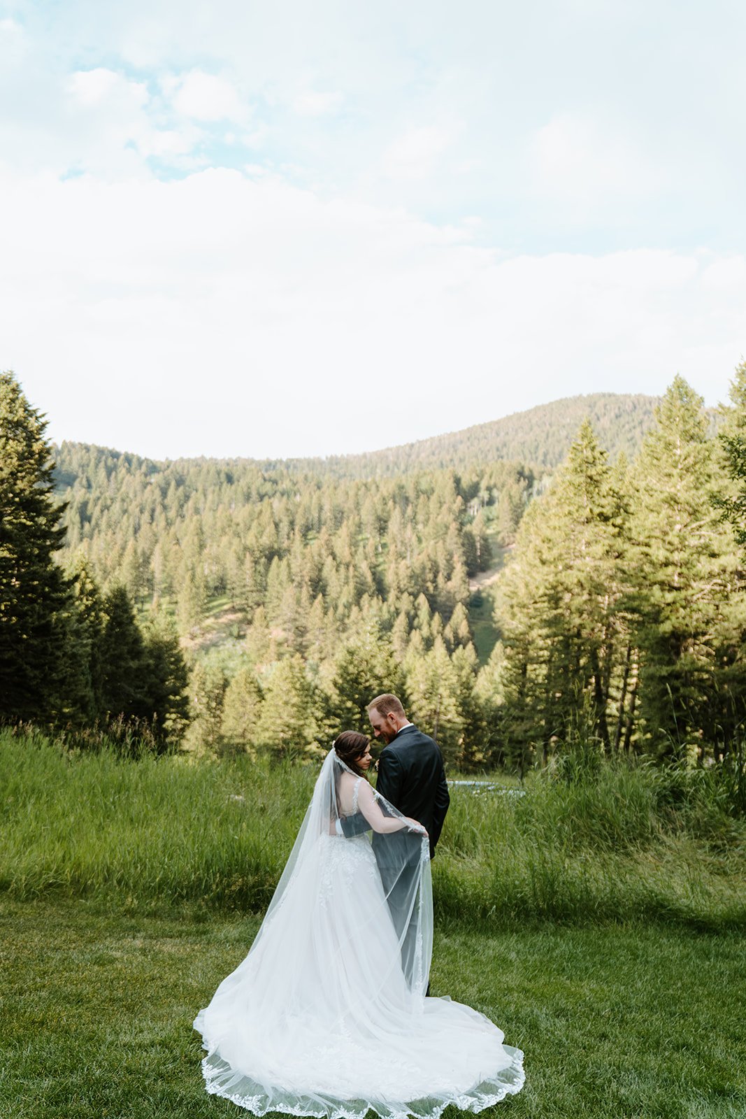 A bride and groom standing close together in a grassy field with a backdrop of forested mountains and a blue sky.
