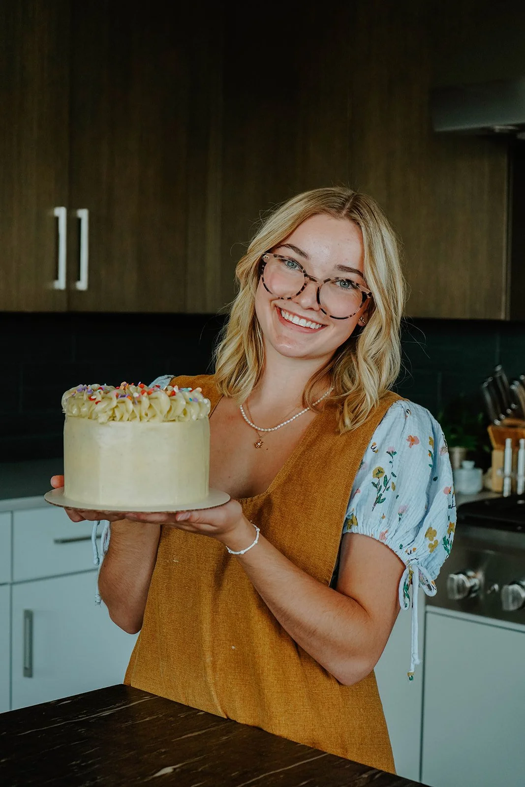A smiling woman with glasses holding a decorated cake in a kitchen.