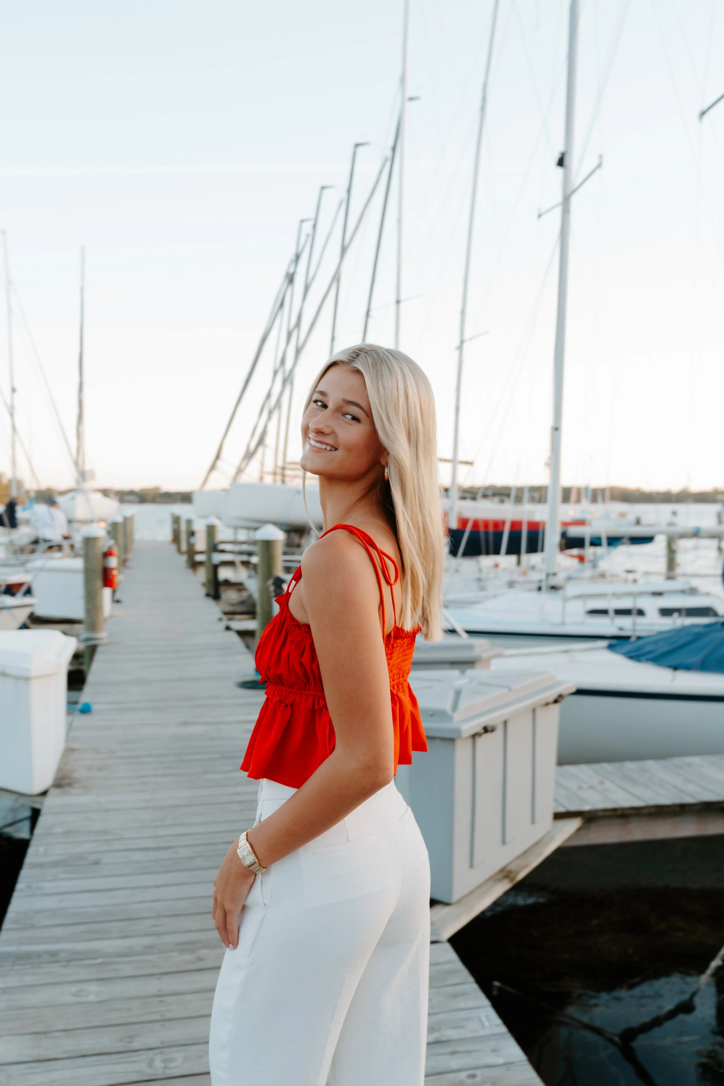 Young woman in a red top and white pants standing on a marina dock with sailboats in the background