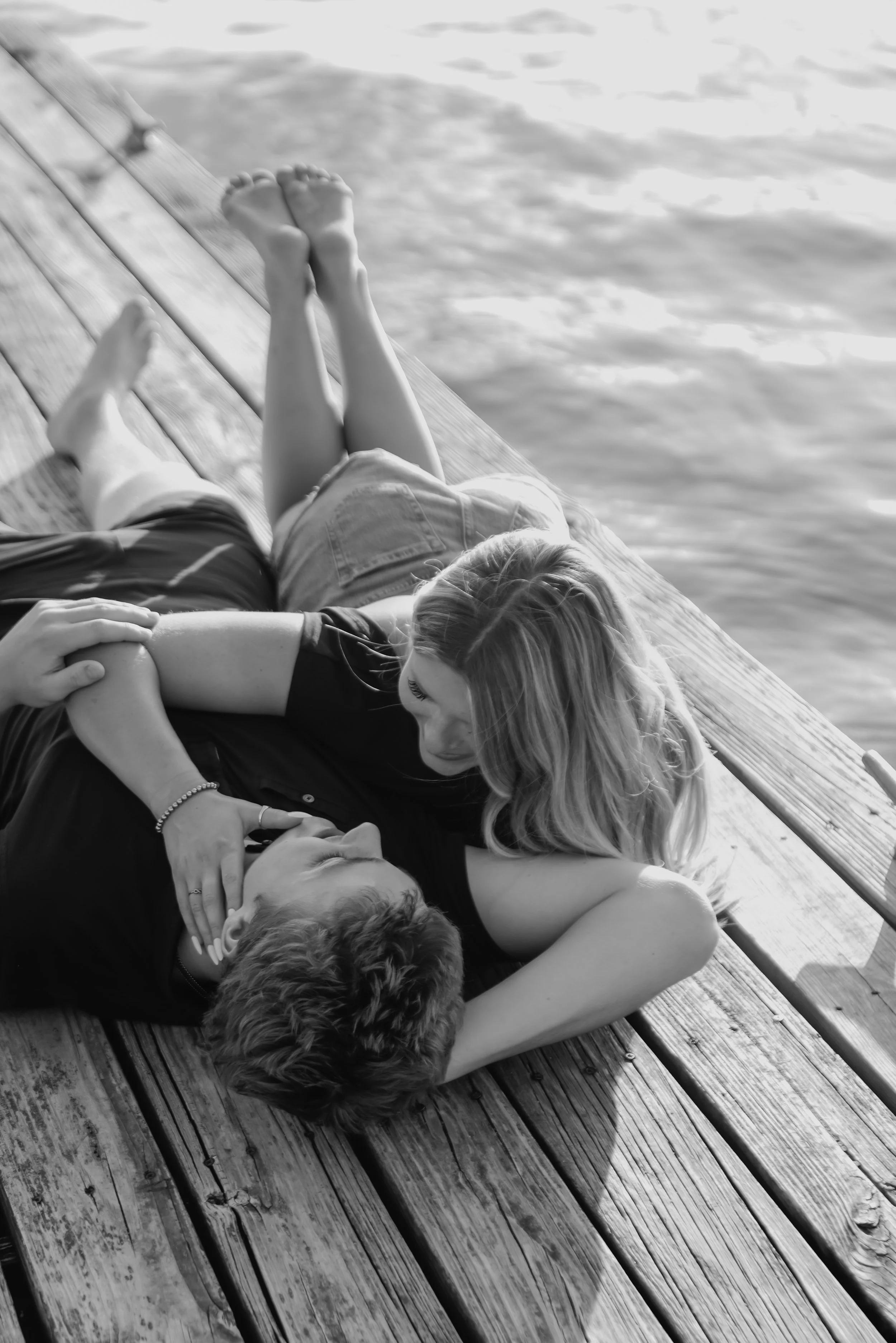A couple lying on a wooden dock by the water, embracing and looking at each other.