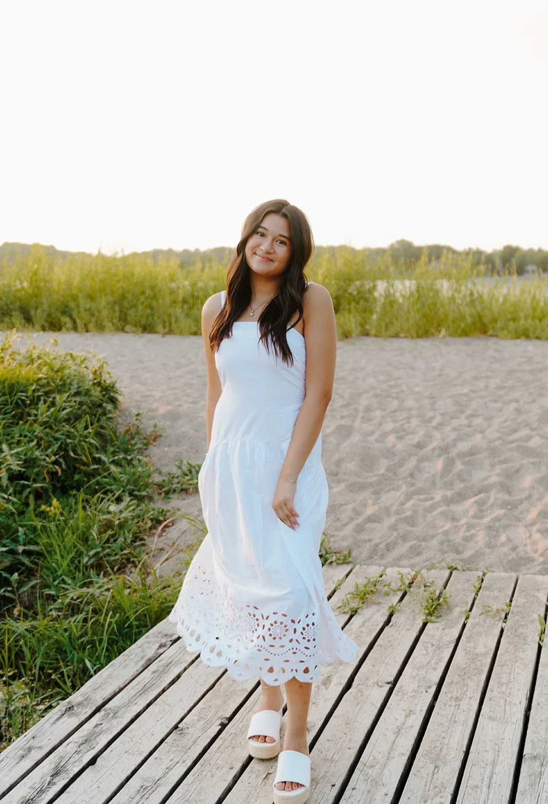 A young woman in a white dress and platform sandals standing on a wooden dock on a sandy area with greenery in the background, smiling at the camera during sunset.