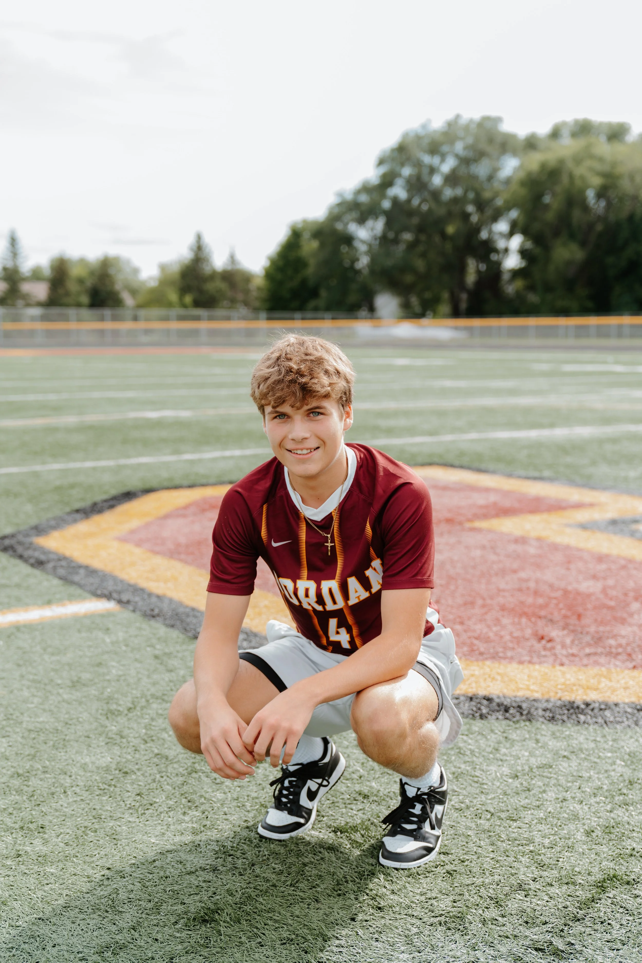 A young male athlete squatting on a sports field, wearing a maroon jersey with the word 'JORDAN' and the number 4, shorts, and sneakers, smiling at the camera.