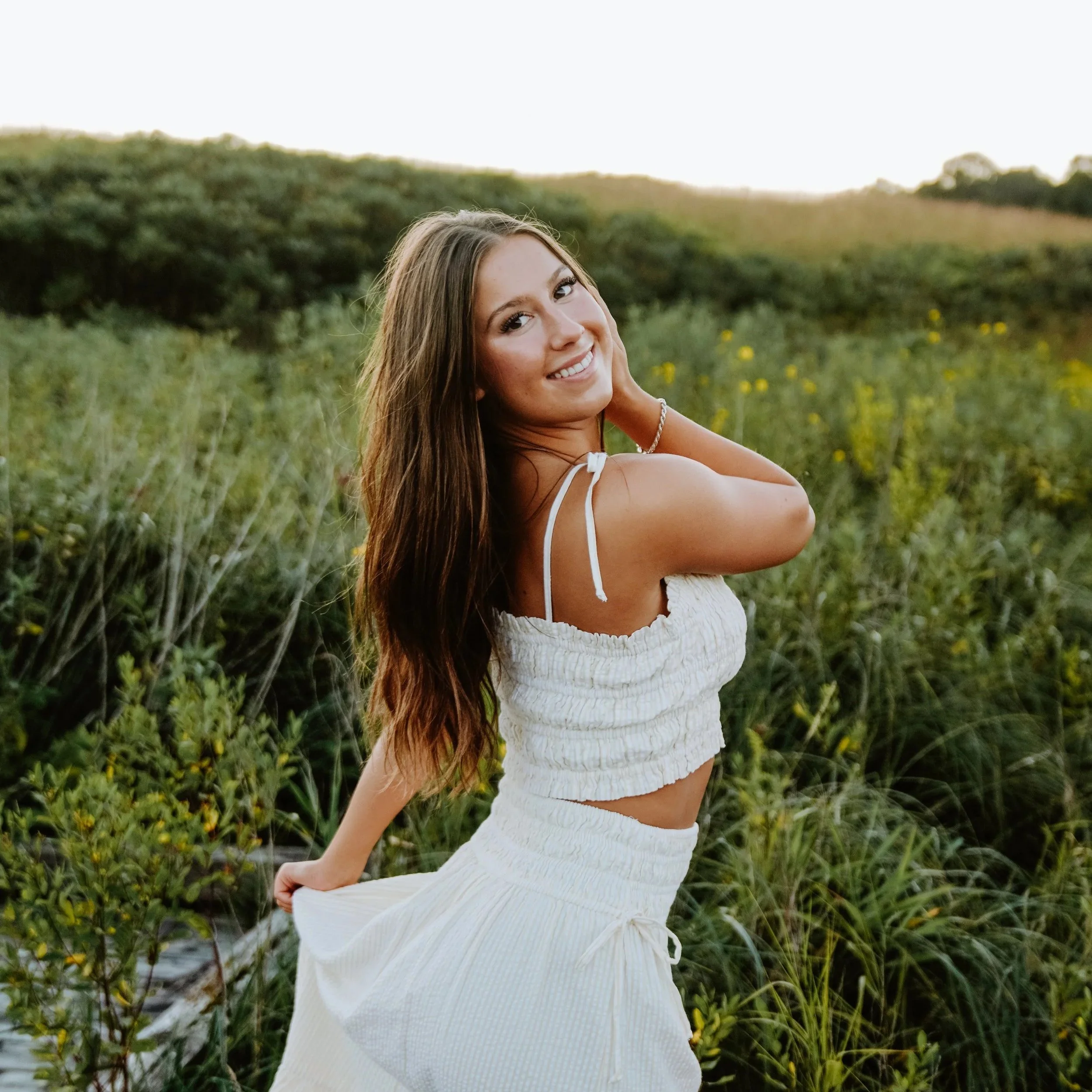 A young woman with long brown hair smiling in a white summer outfit in a green outdoor field during sunset.