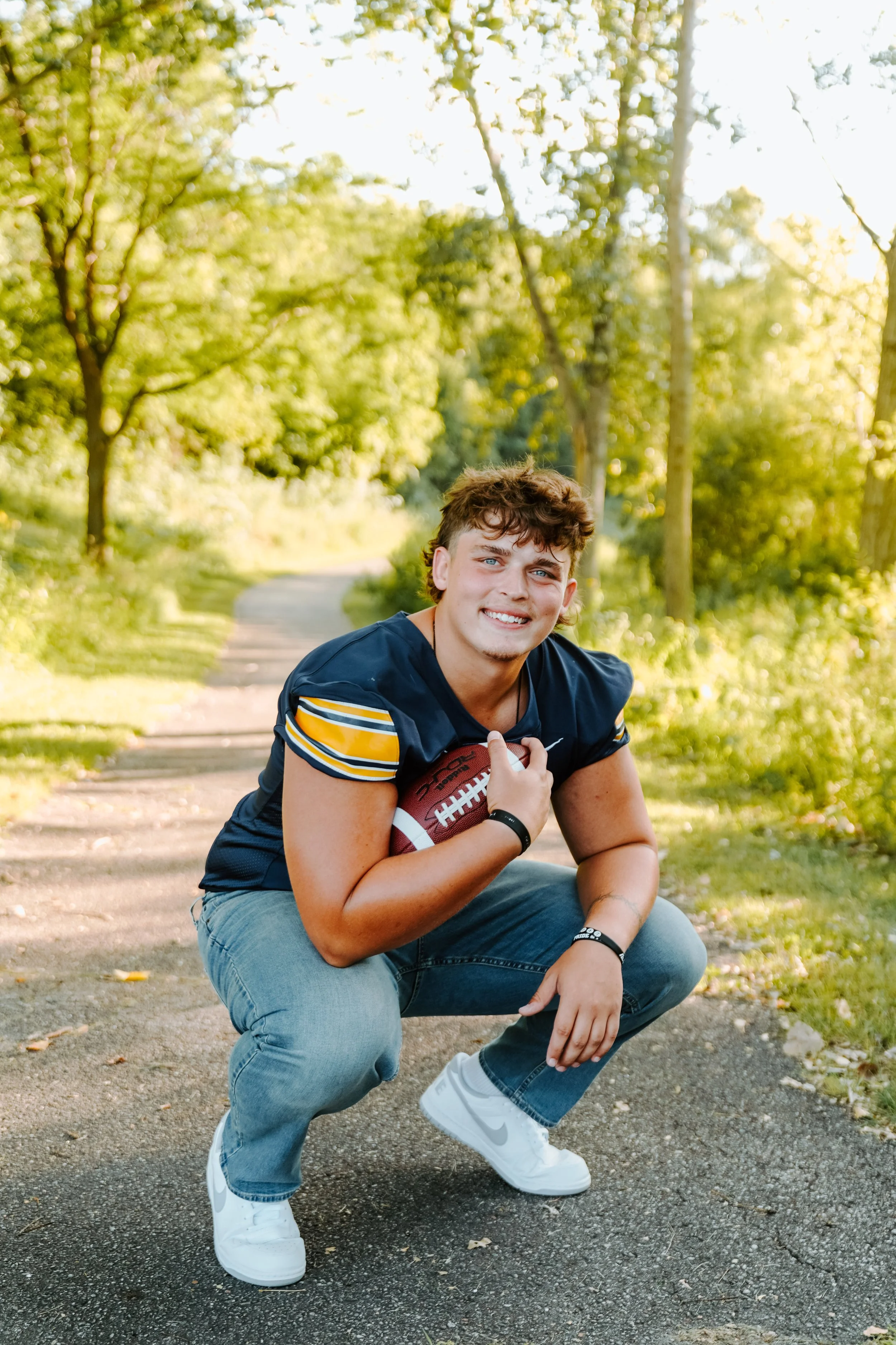 A young man crouching on a paved trail, holding a football, smiling at the camera outdoors surrounded by trees in sunlight.