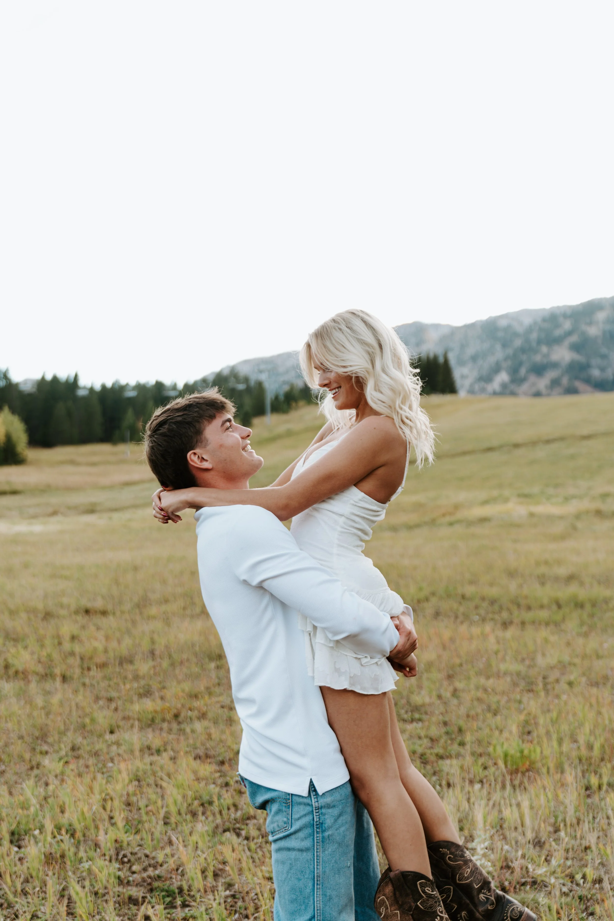 A smiling young man in a white shirt and jeans lifts a woman in a white dress with blonde hair in a field with mountains in the background.