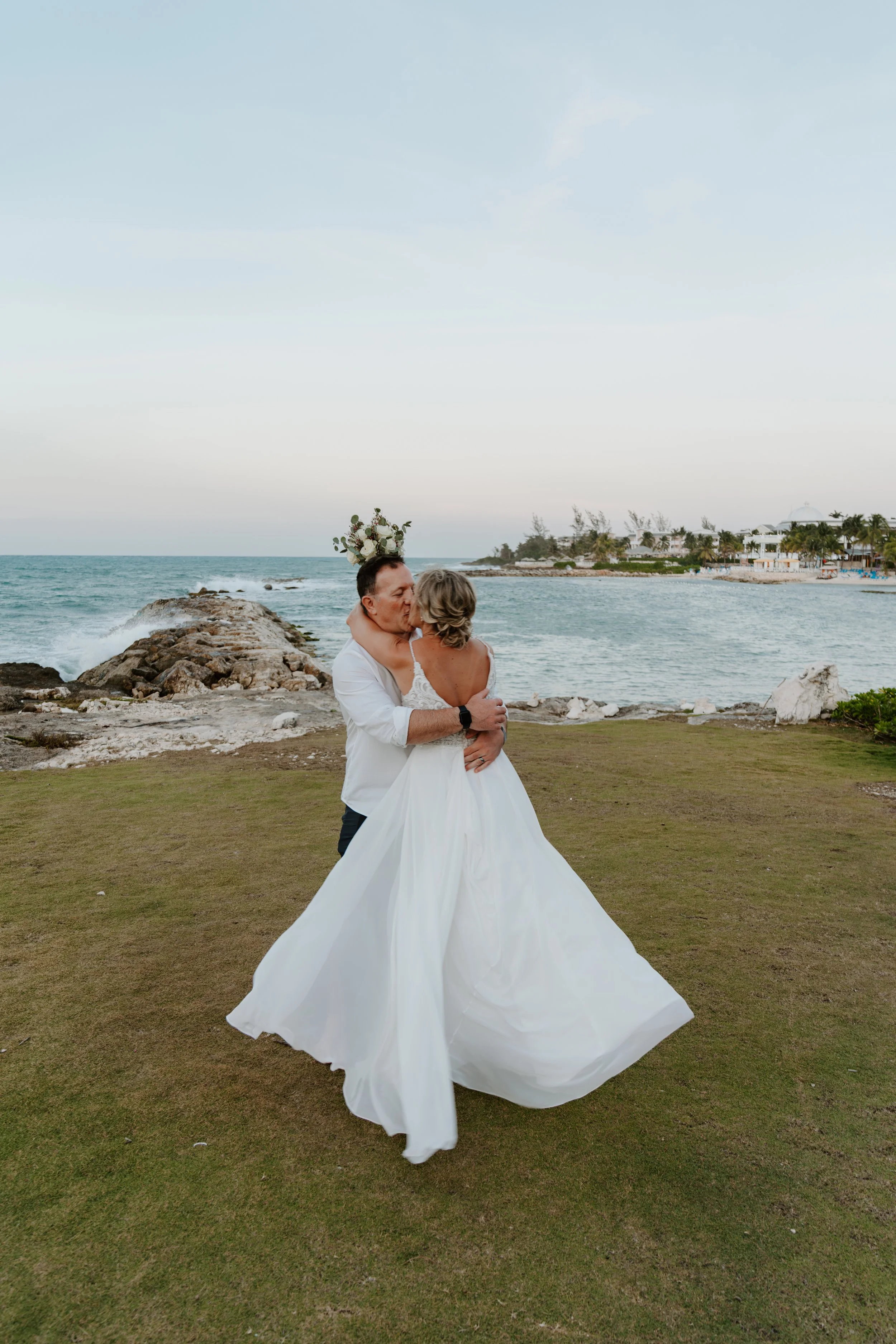A newlywed couple sharing a kiss on the beach, with the ocean, rocks, and distant buildings in the background, during sunset.
