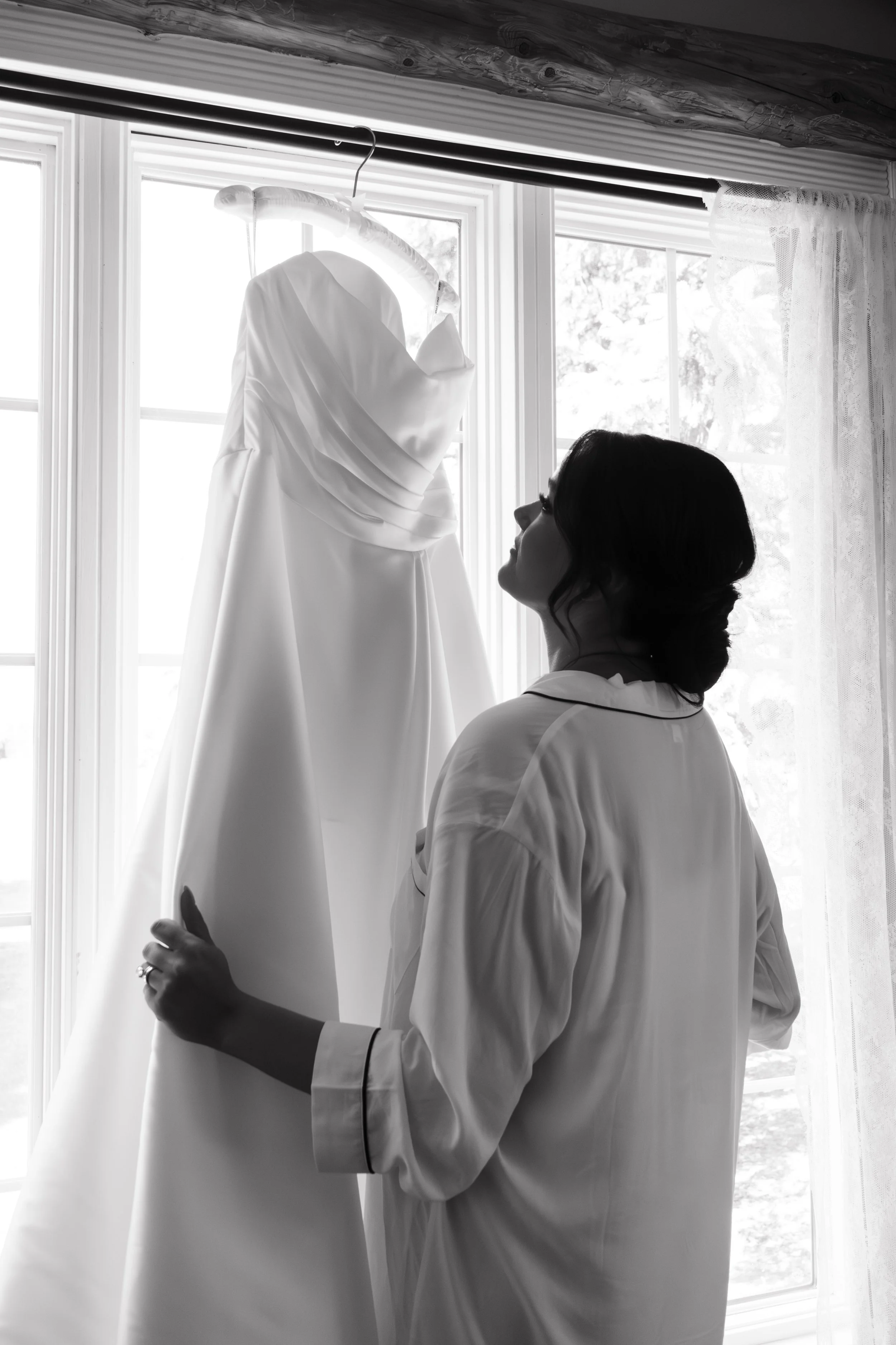 A woman in silk pajamas looks at a wedding dress hanging in front of a window.