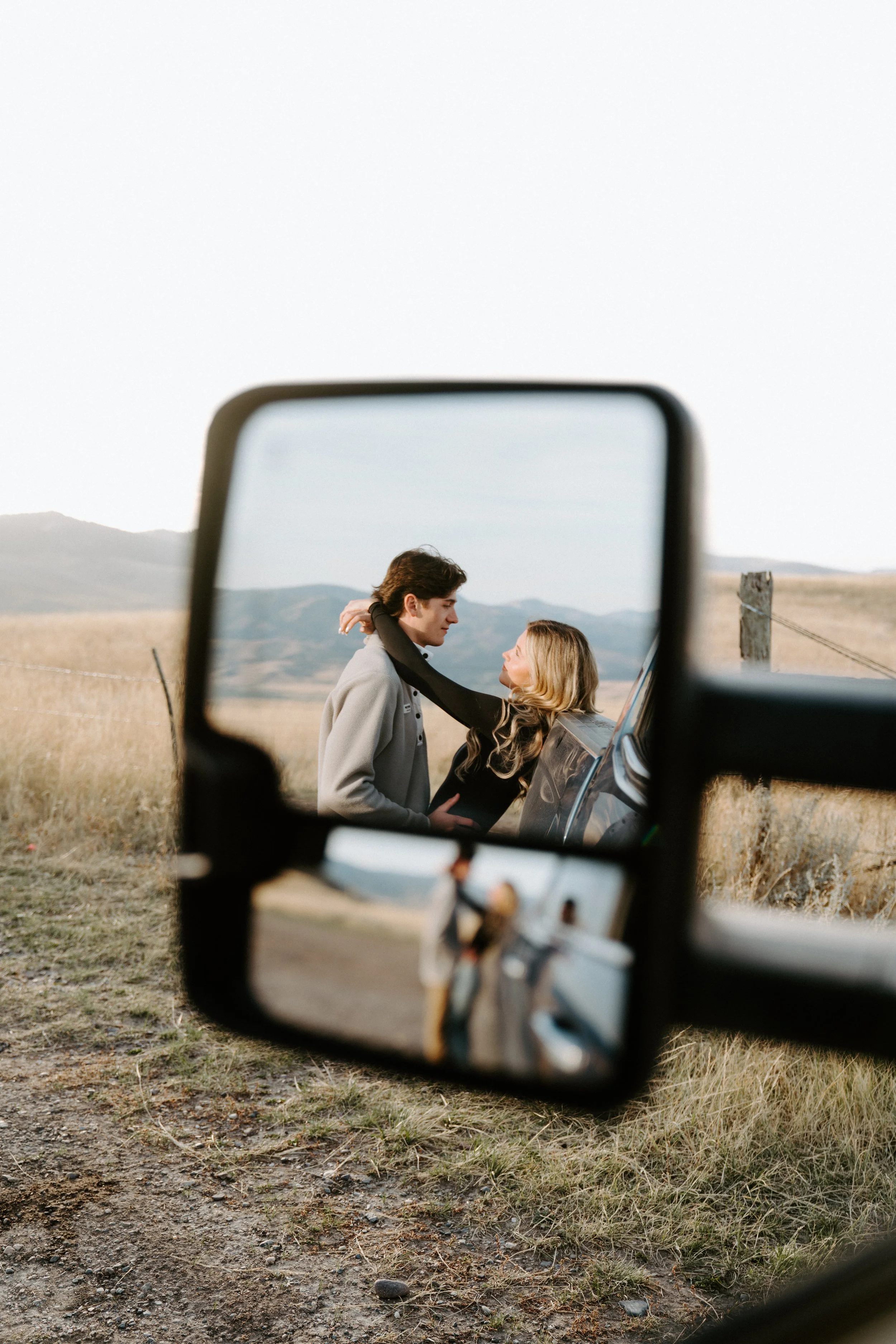 A romantic moment between a man and woman captured in the side mirror of a vehicle, with open countryside and hills in the background.