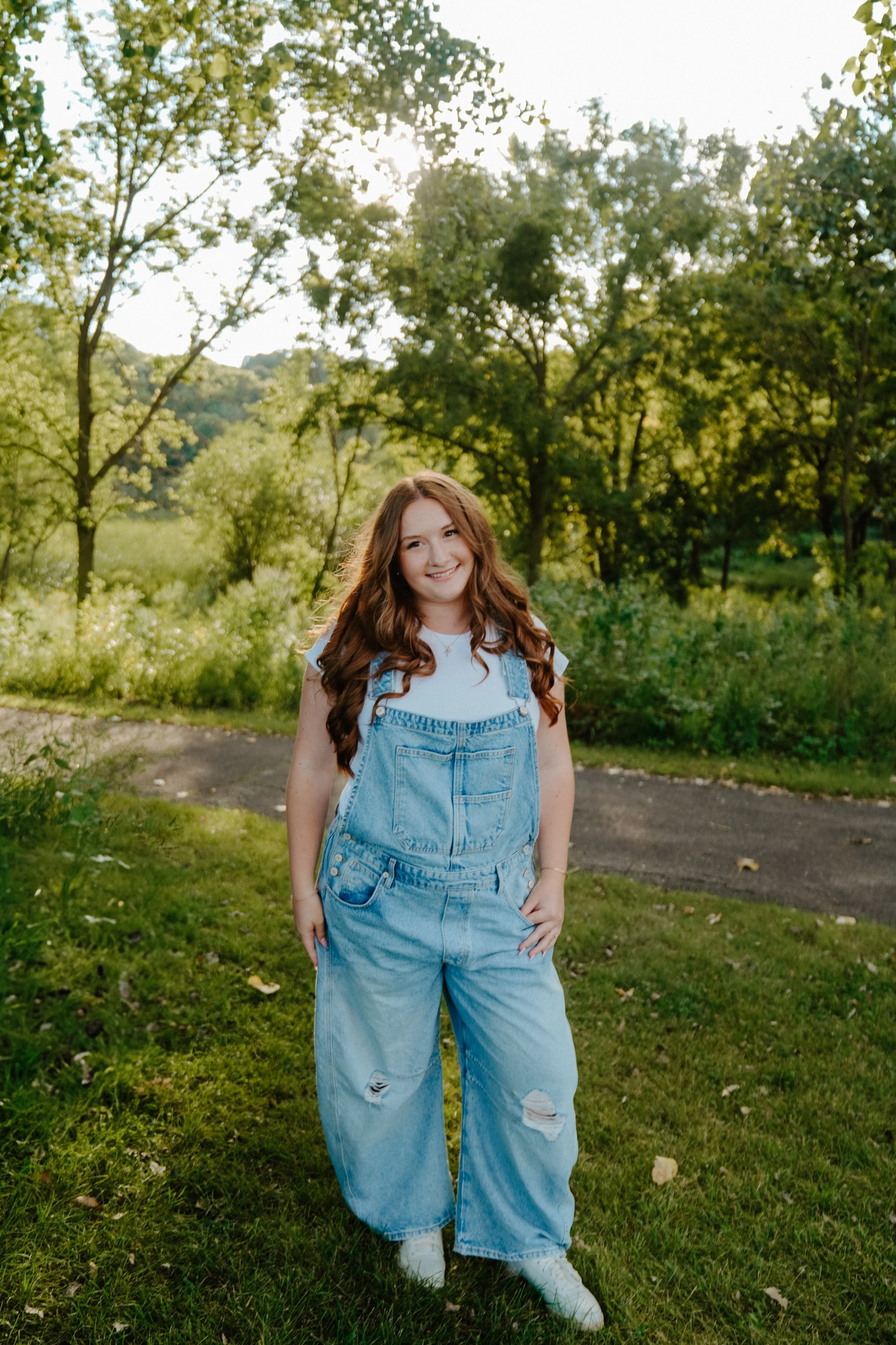 A young woman with long, wavy brown hair is standing outdoors on a grassy path, smiling at the camera. She is wearing a white t-shirt, light blue ripped denim overalls, and white sneakers. The background features green trees and shrubs under sunlight