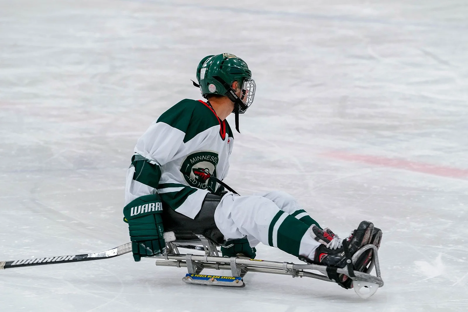 A hockey player in a Minnesota Wild jersey sitting on the ice in a sled hockey chair, wearing a helmet and protective gear.