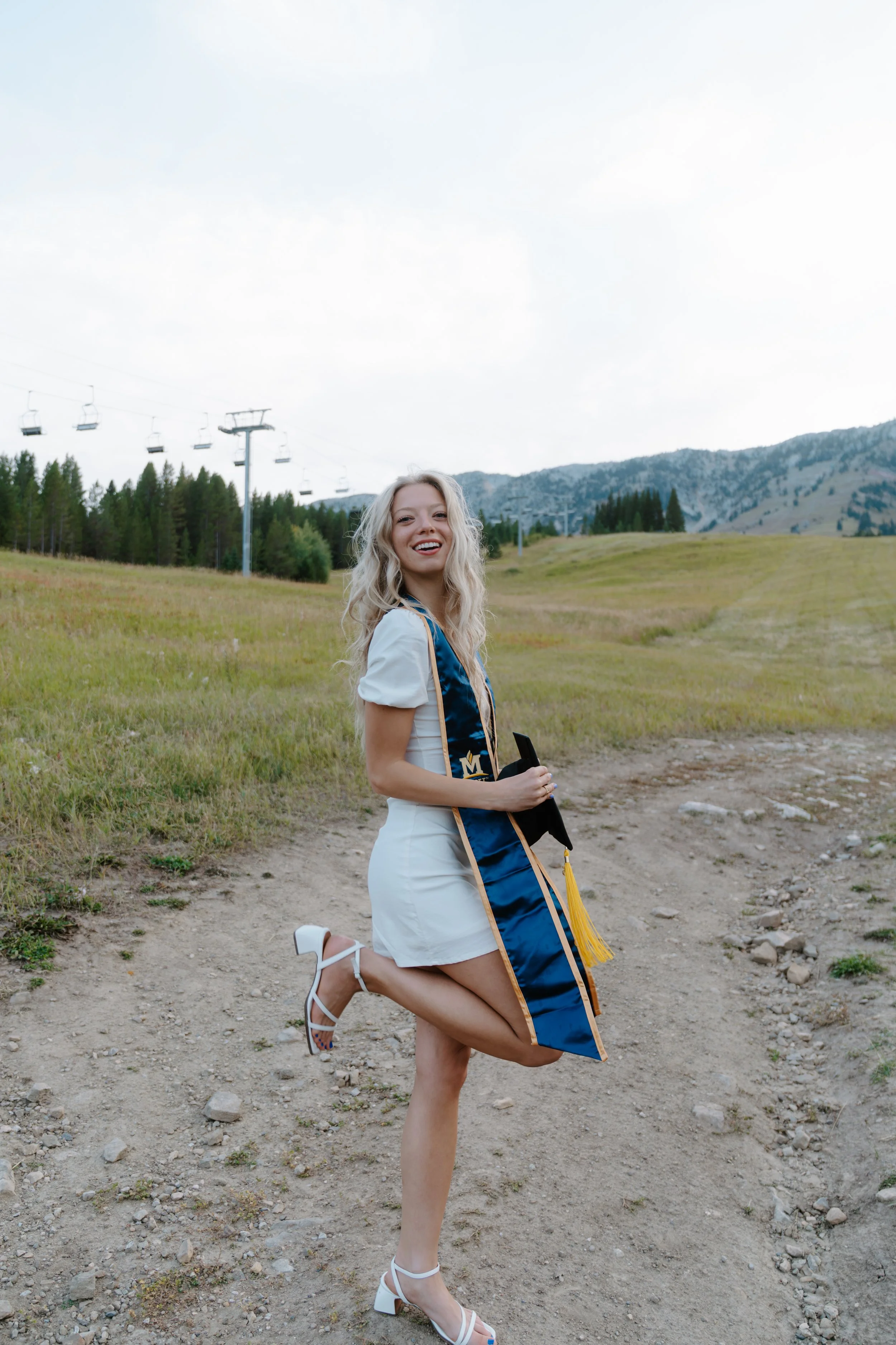 A young woman celebrating graduation outdoors on a dirt trail, wearing a white dress and heels, holding her diploma, with a scenic mountain and forest background.