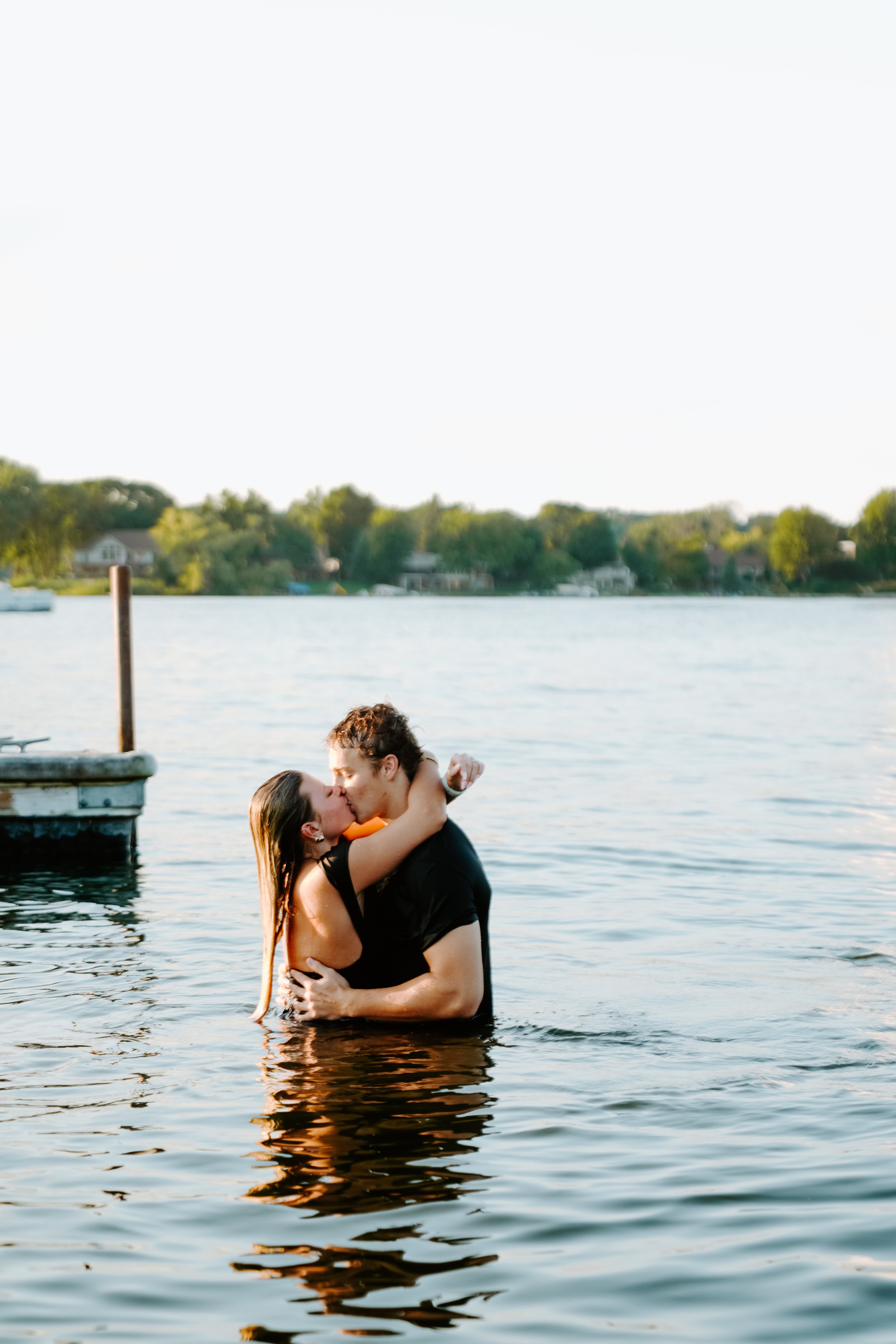 A couple embraces and kisses in a river or lake during daylight, with trees and houses in the background.