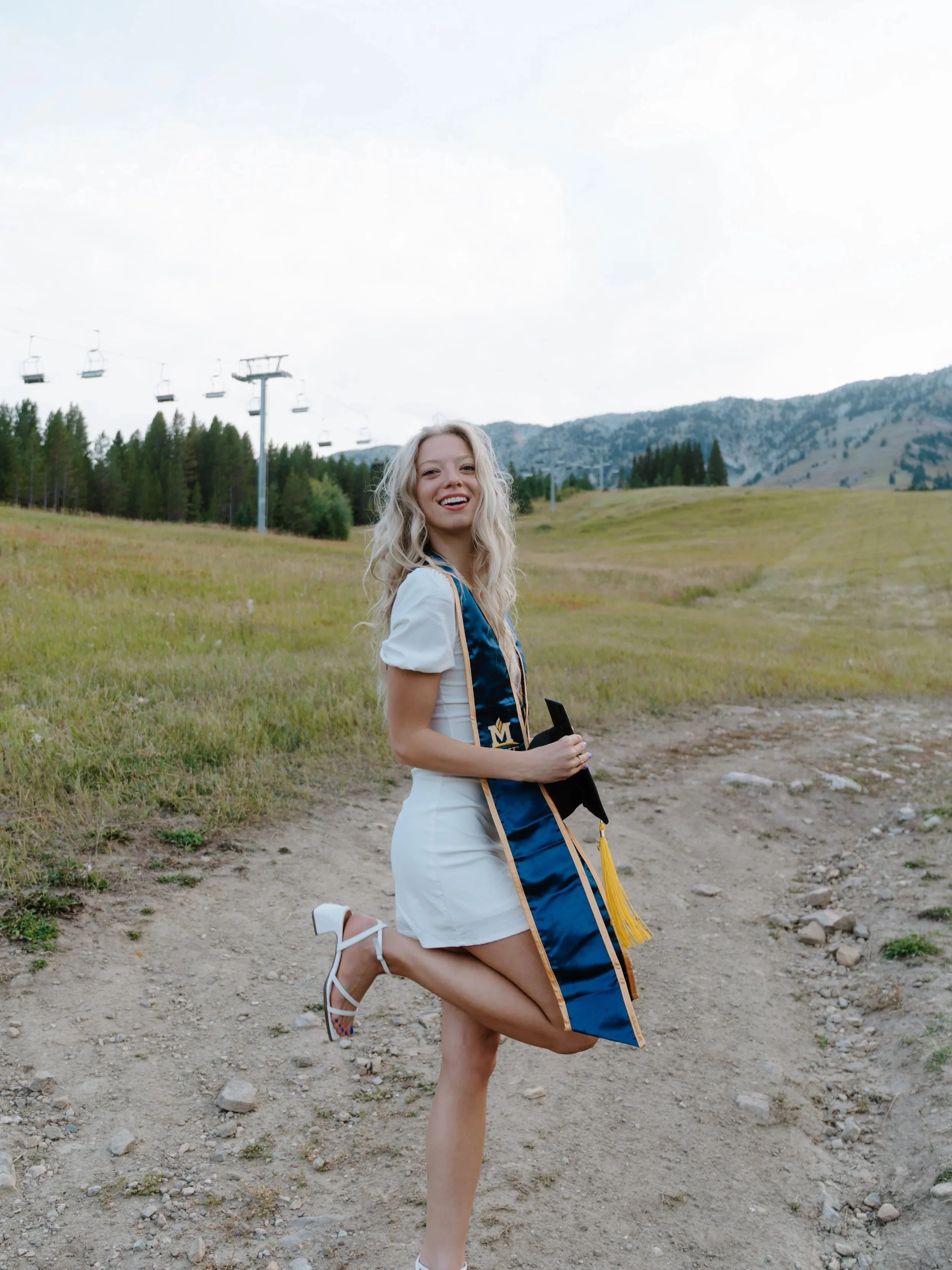 A young woman celebrating graduation outdoors, wearing a white dress and white heels with an academic stole, standing on a dirt path in a grassy field with mountains and ski lifts in the background, smiling and posing with one leg lifted.