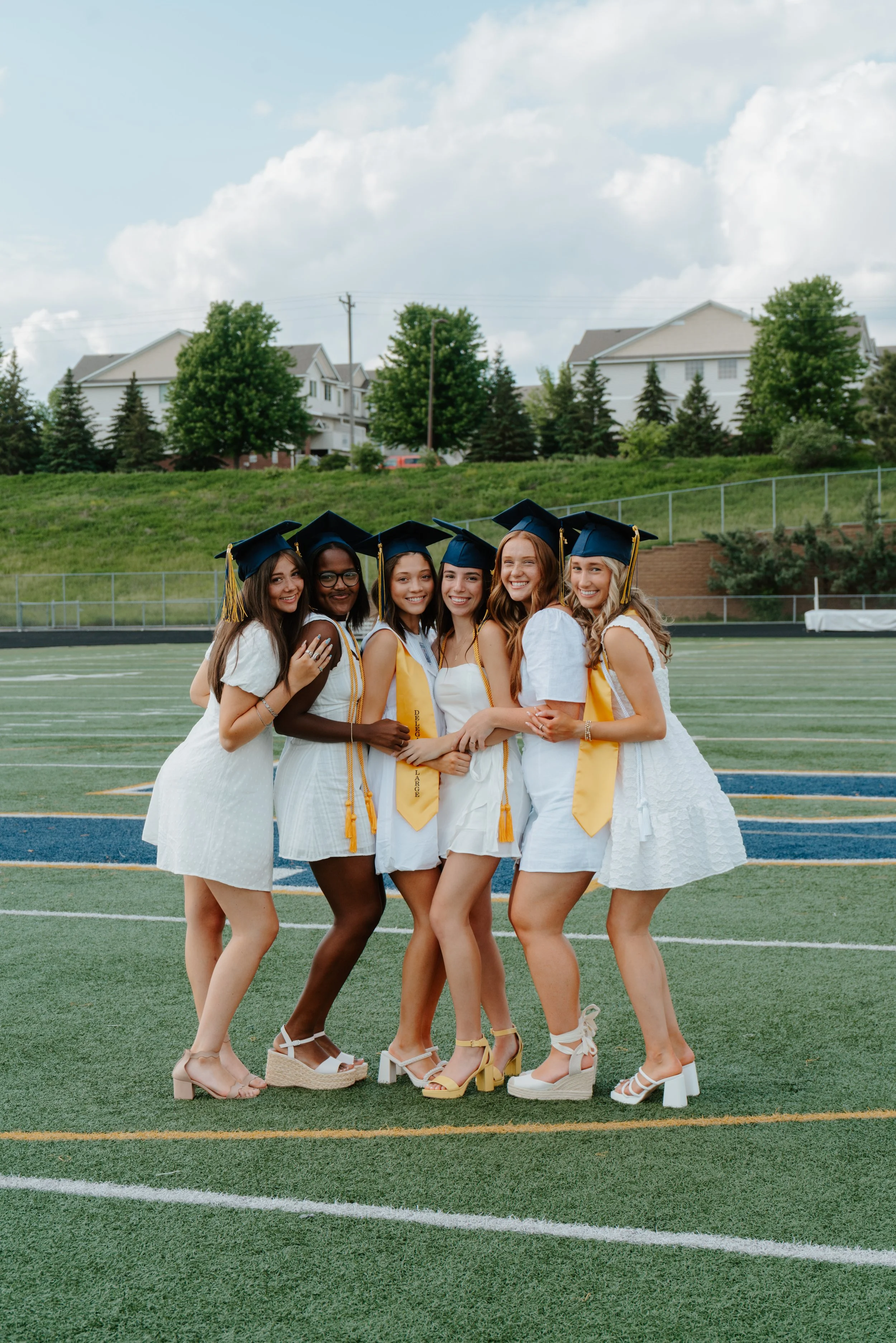 Group of six young women in graduation caps and gowns standing on a football field, smiling and hugging each other.