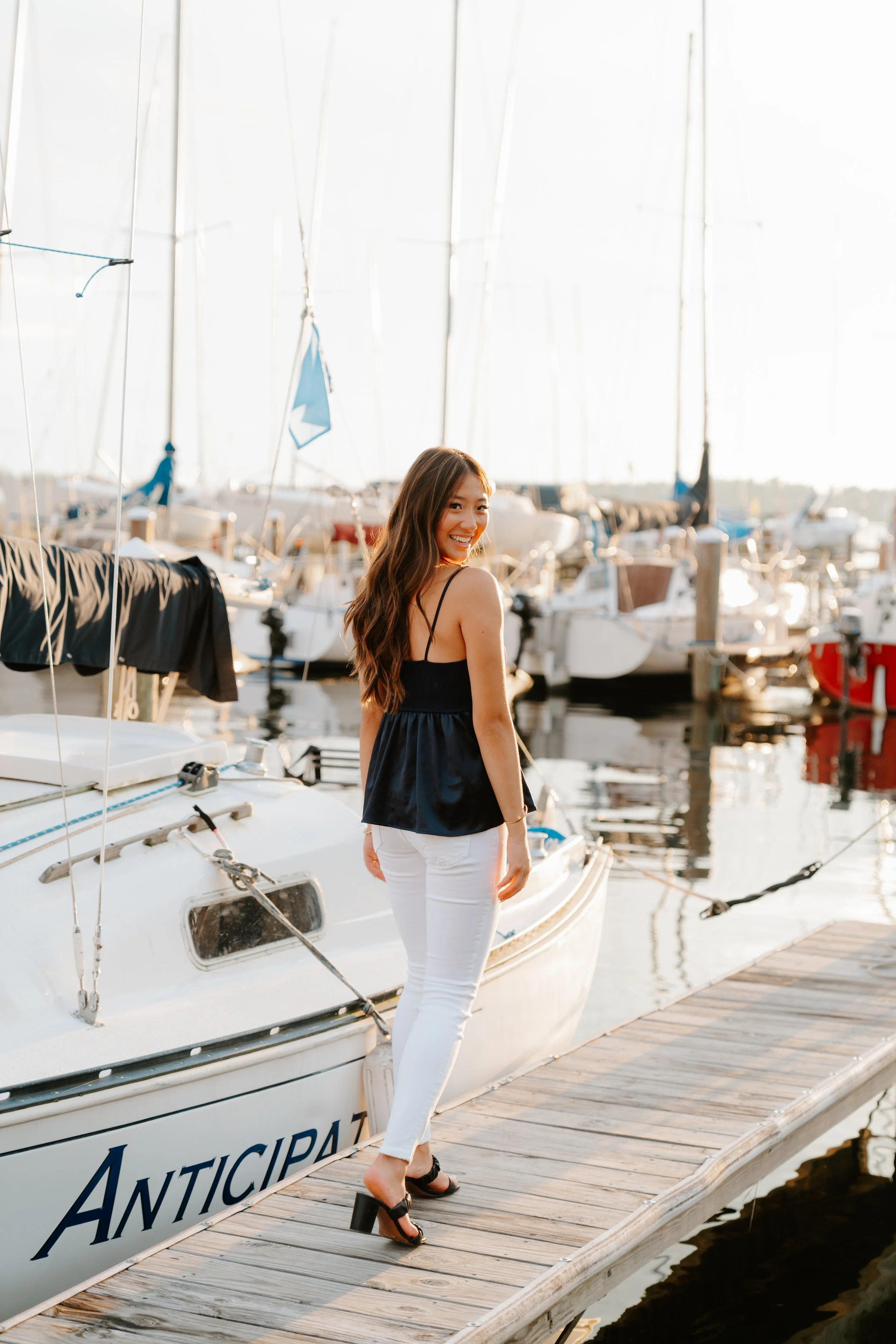 A woman on a wooden dock at a marina, smiling and looking back. She is wearing a black top, white pants, and black high-heeled sandals. Several sailboats are docked in the background.