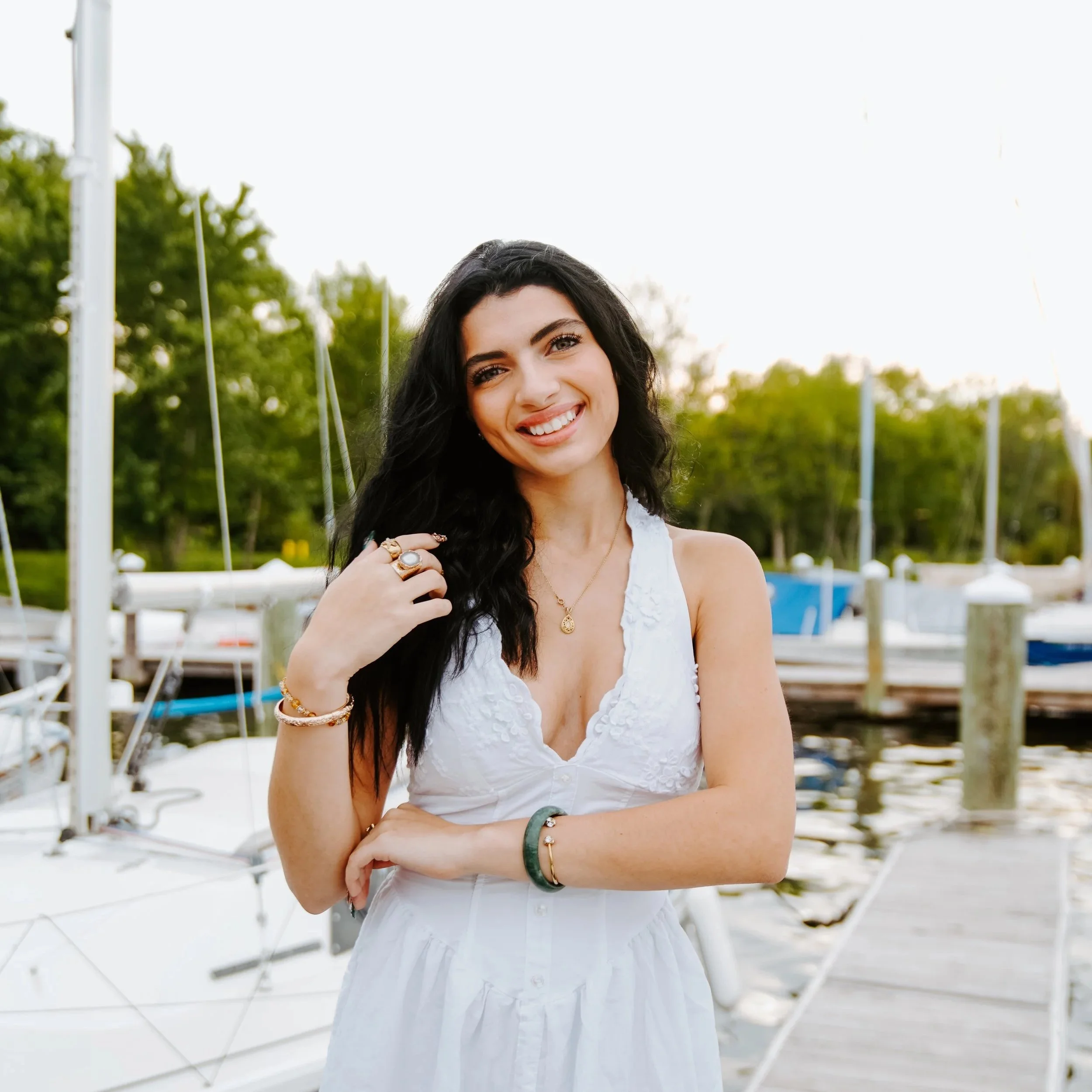 A woman with long black hair, wearing a white sleeveless dress, smiling at the camera at a marina with sailboats and green trees in the background.