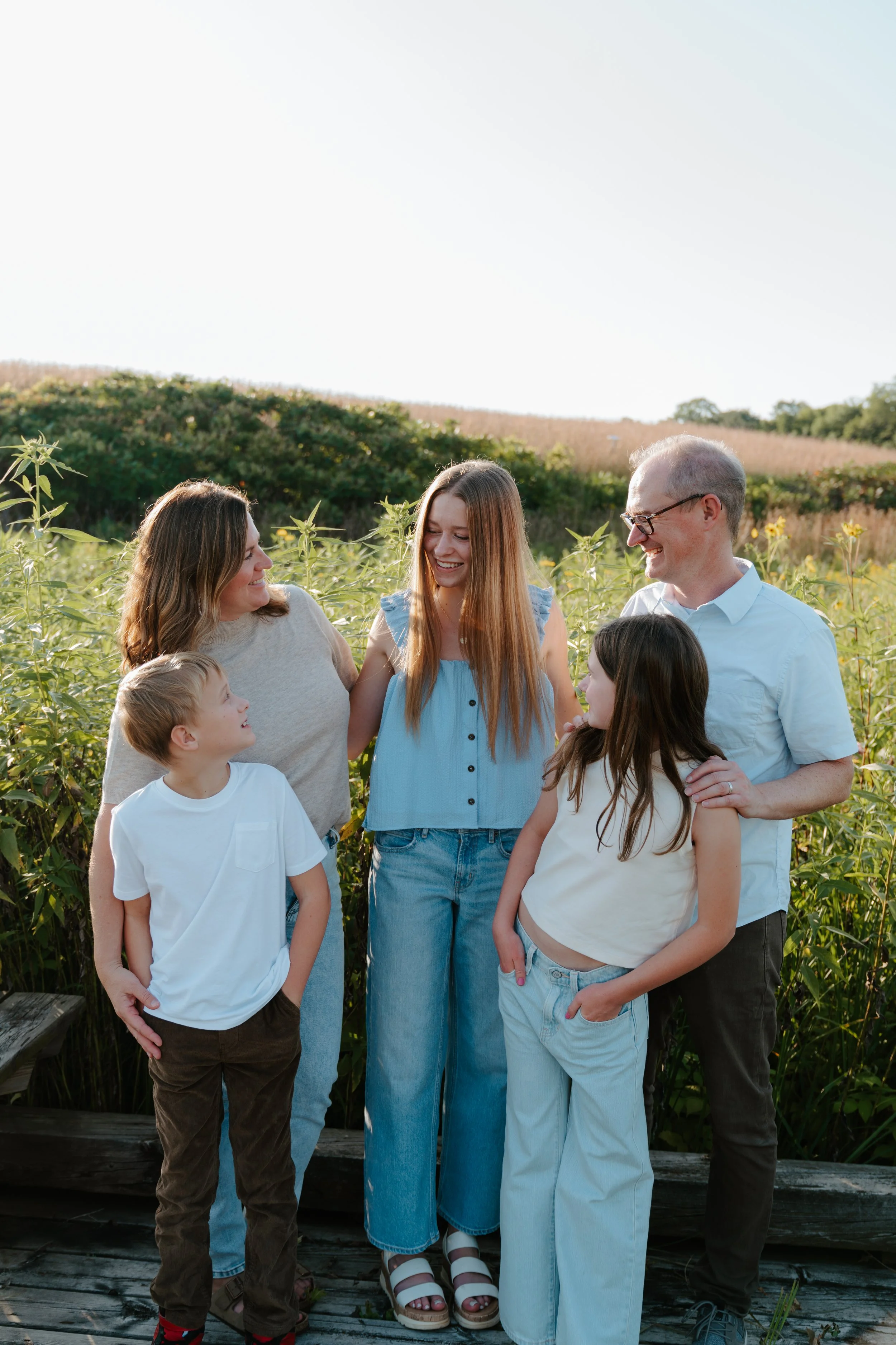 Family of six standing outdoors in a field of tall plants, smiling and looking at each other, with a hill in the background on a sunny day.