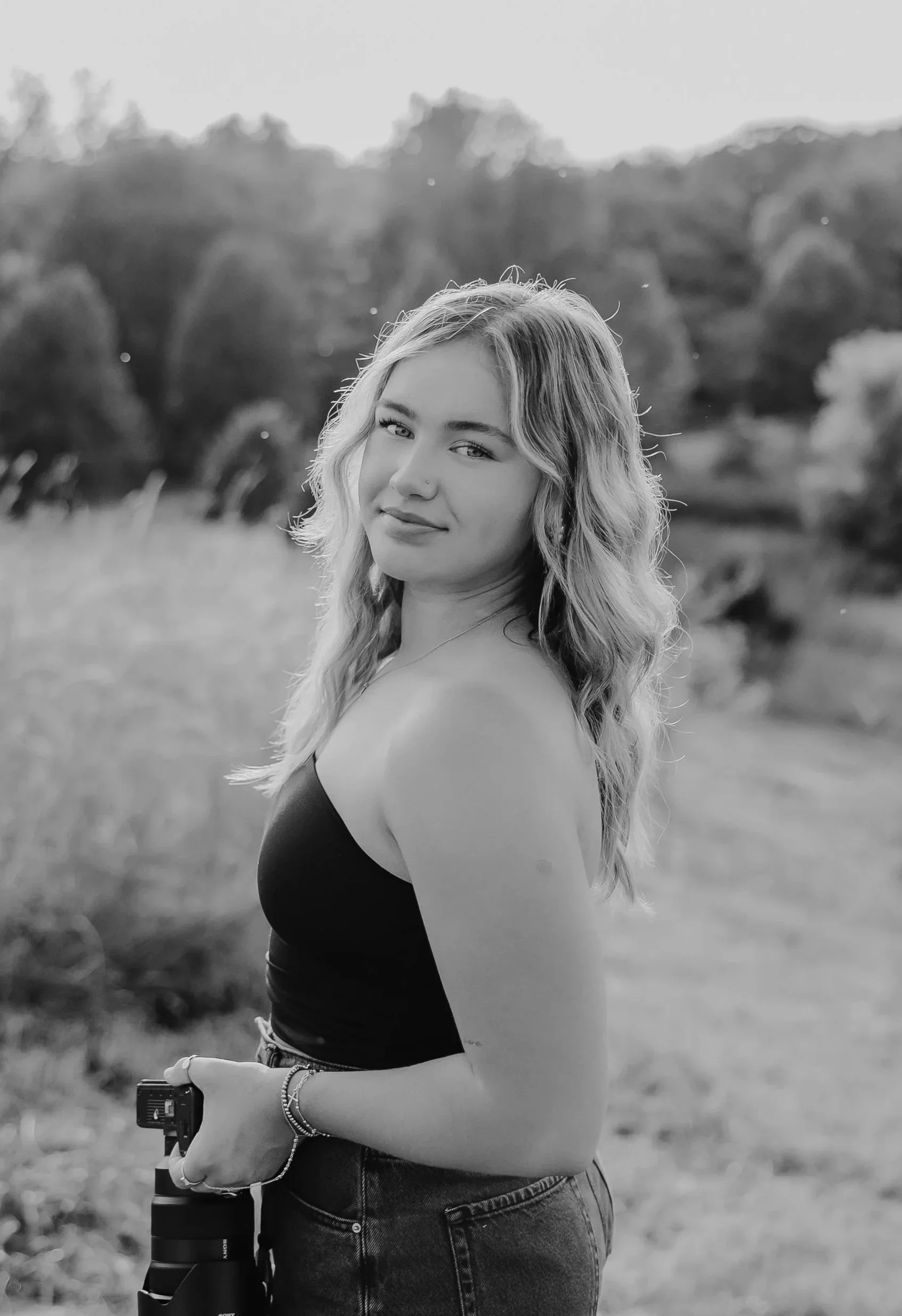 Black and white photo of a young woman with long wavy hair standing outdoors, holding a camera in her right hand, and looking at the camera with a slight smile, with trees and open landscape in the background.