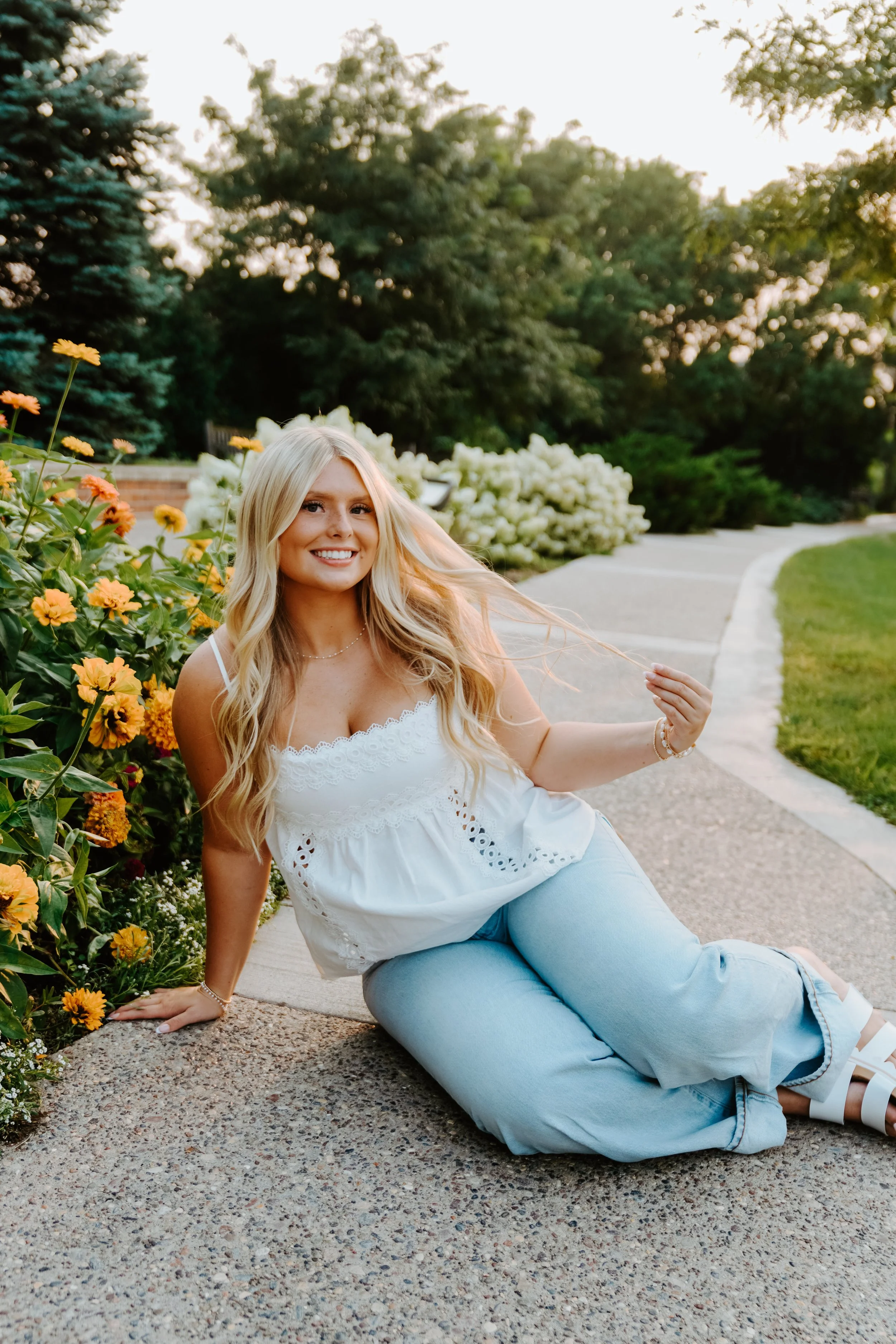 A young woman with long blonde hair, smiling, sitting on a sidewalk next to a flower bed with orange and yellow flowers, in a park-like setting with trees and greenery in the background during sunset.