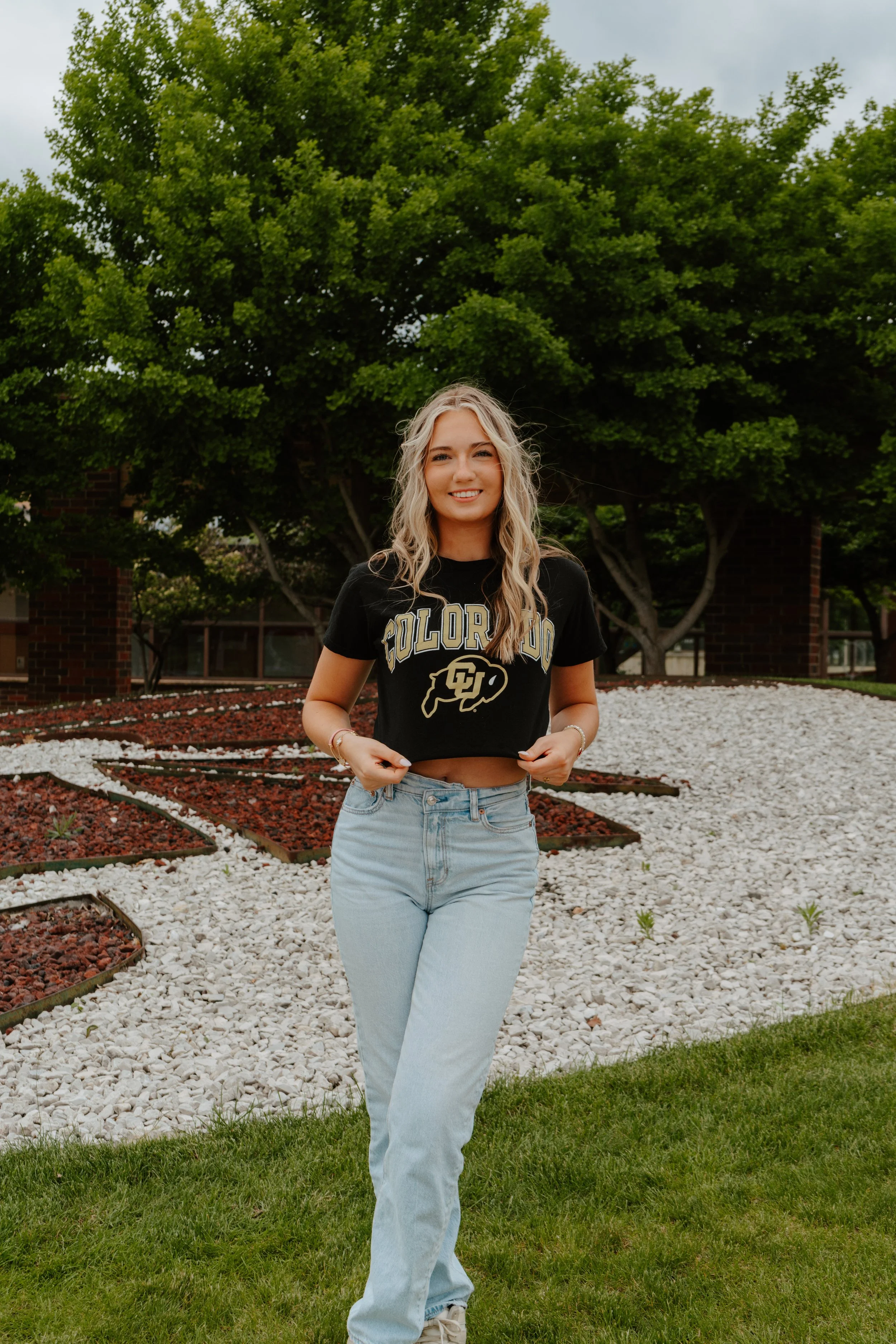 A young woman standing outdoors on a grassy area with a large green tree behind her, wearing a black Colorado University t-shirt and light blue jeans, smiling at the camera.